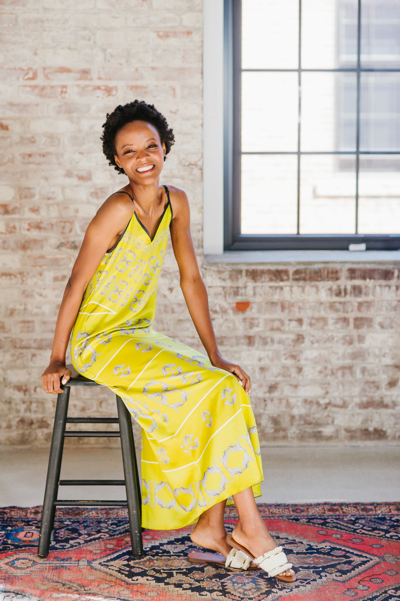 A woman sitting on a black wooden stool, smiling, in a room with a brick wall and large window, wearing a yellow patterned dress and white sandals.