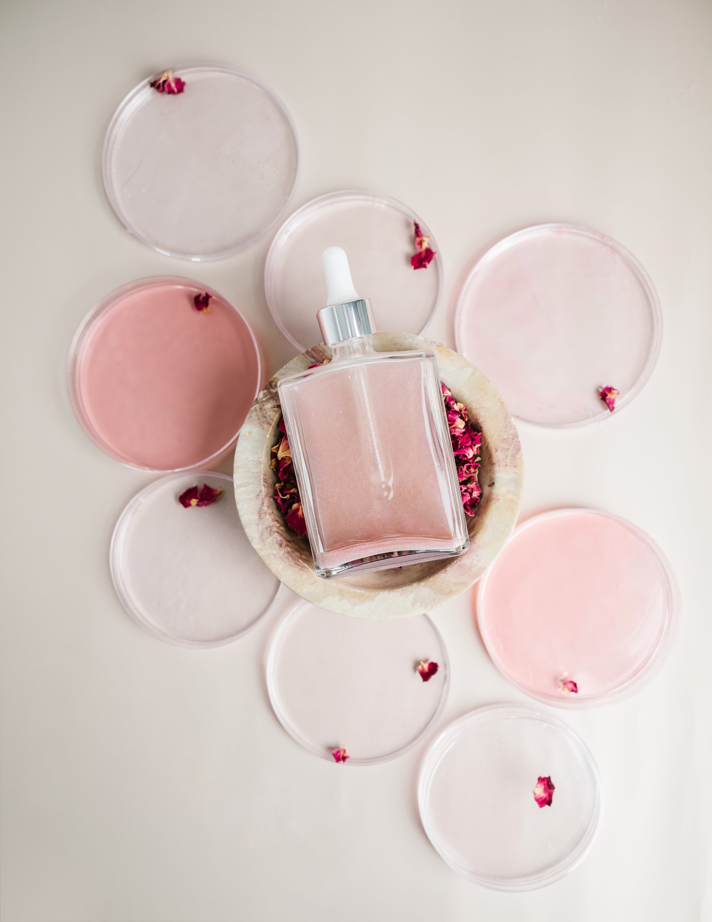 A glass bottle with a dropper cap containing pink liquid, surrounded by circular petri dishes with pink and clear substances, and scattered dried flower petals on a white surface.