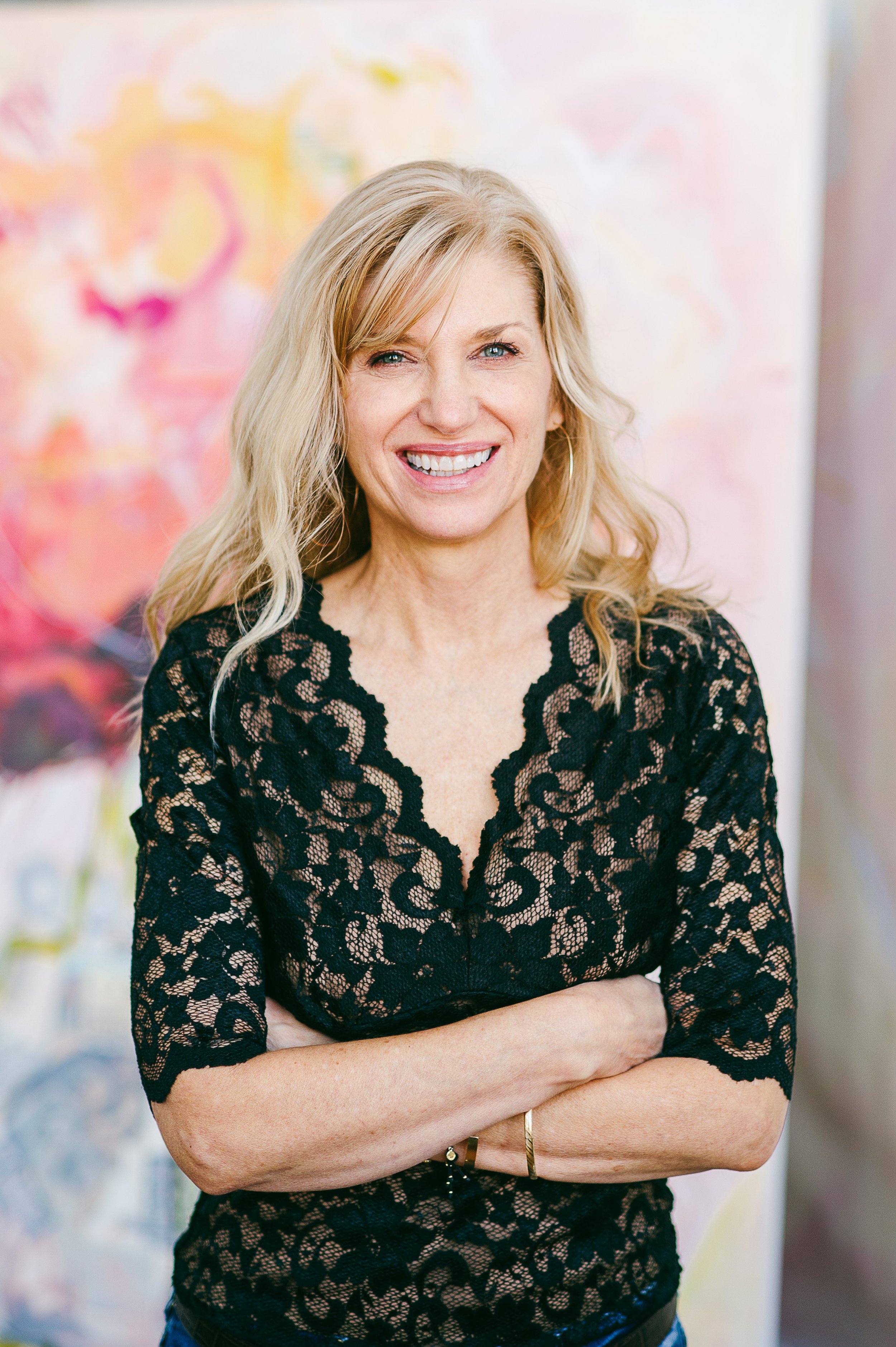 A smiling woman with wavy blonde hair, wearing a black lace top, standing with arms crossed against an abstract colorful background.