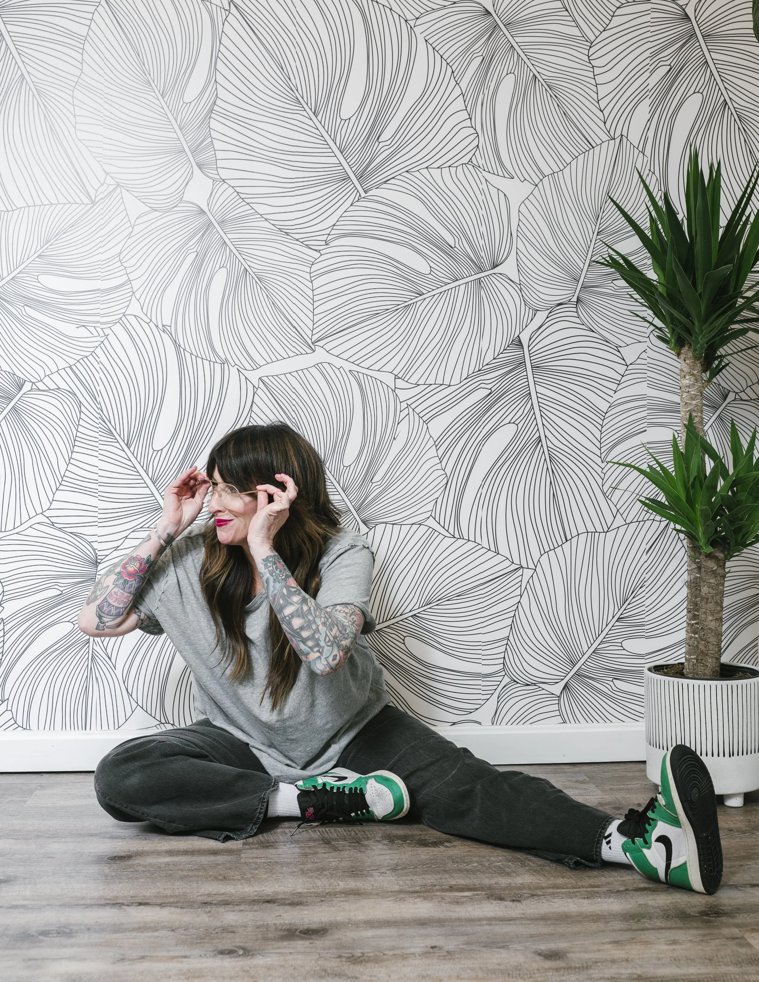 Branding photography featuring a woman with tattoos on her arms, wearing glasses, a gray t-shirt, black pants, and green and white sneakers, sitting on the floor in front of a wall with a black and white leaf pattern, adjusting her glasses. 