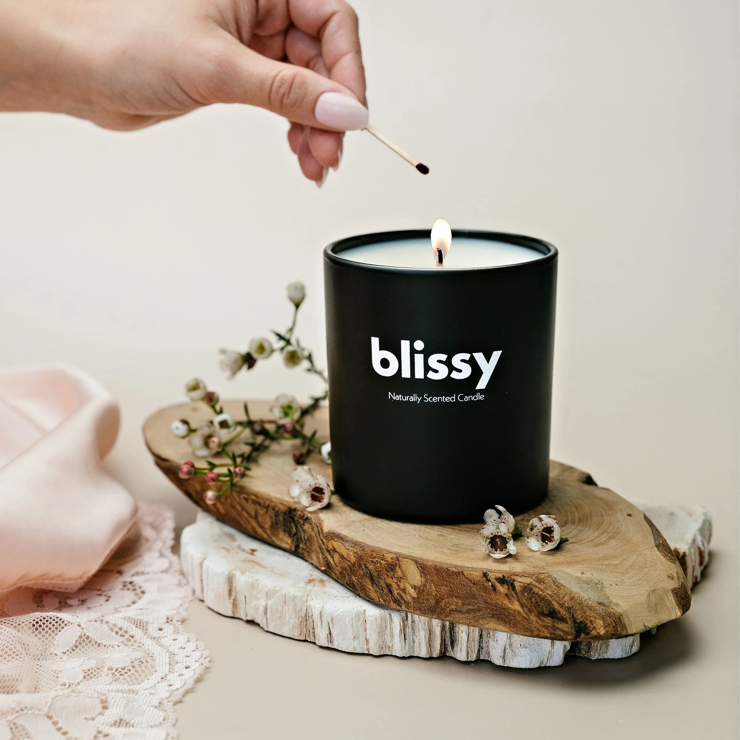 Product photography featuring a person lighting a black candle labeled 'blissy' on a wooden tray surrounded by small white and pink flowers, with a pink cloth and lace fabric nearby.