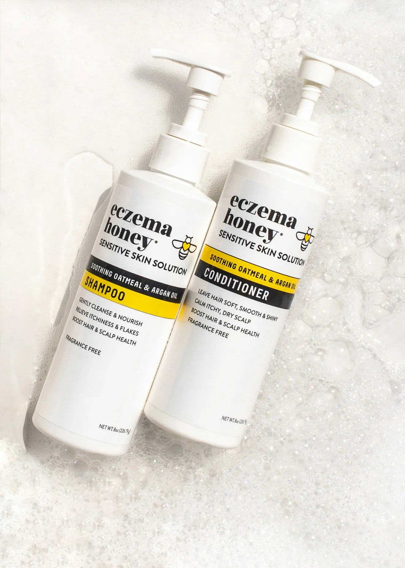 Two white bottles of Eczema Honey skincare products, one shampoo and one conditioner, placed on a bubbly soap background.