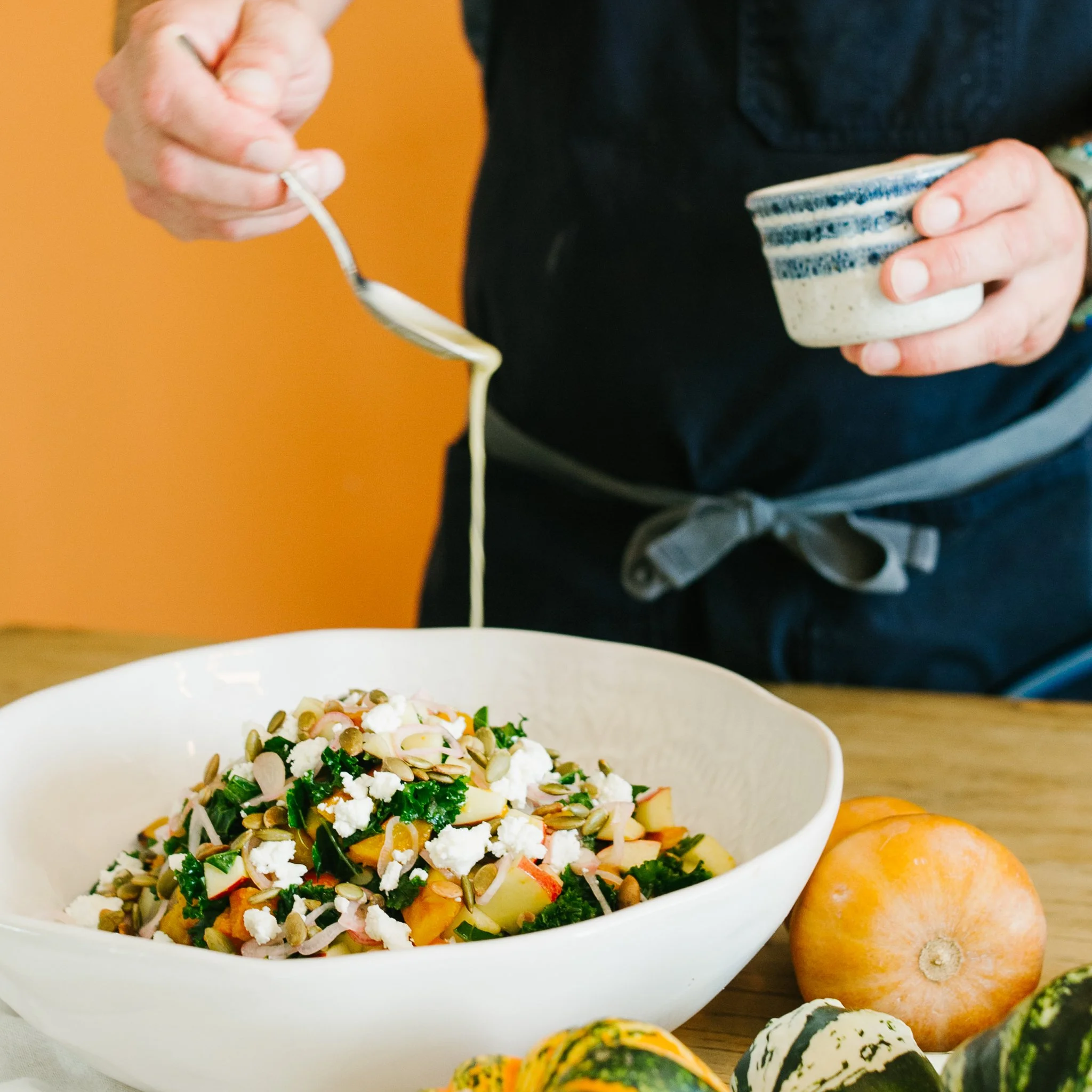 A person is pouring dressing or sauce onto a colorful salad in a white bowl, with a pumpkin and gourds on the table nearby.