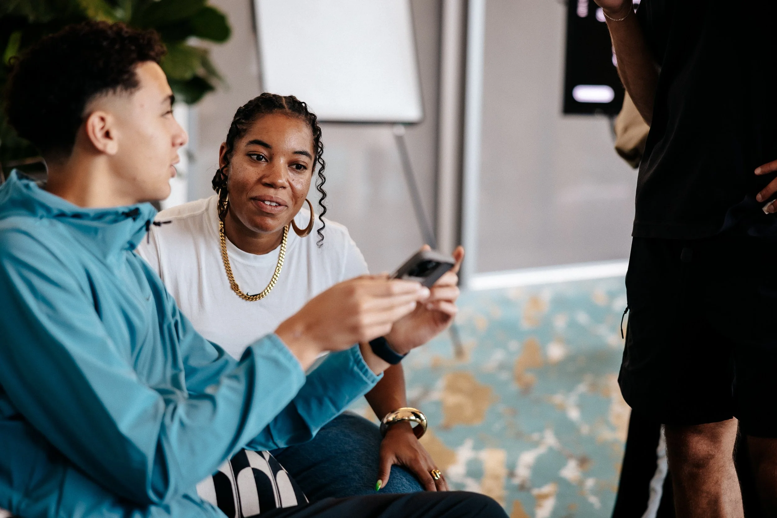 Two women are seated and looking at a smartphone, engaging in a conversation. One woman has curly hair, is wearing jewelry, and a white t-shirt, while the other has short hair and is wearing a blue jacket. A third person stands nearby, partially visi