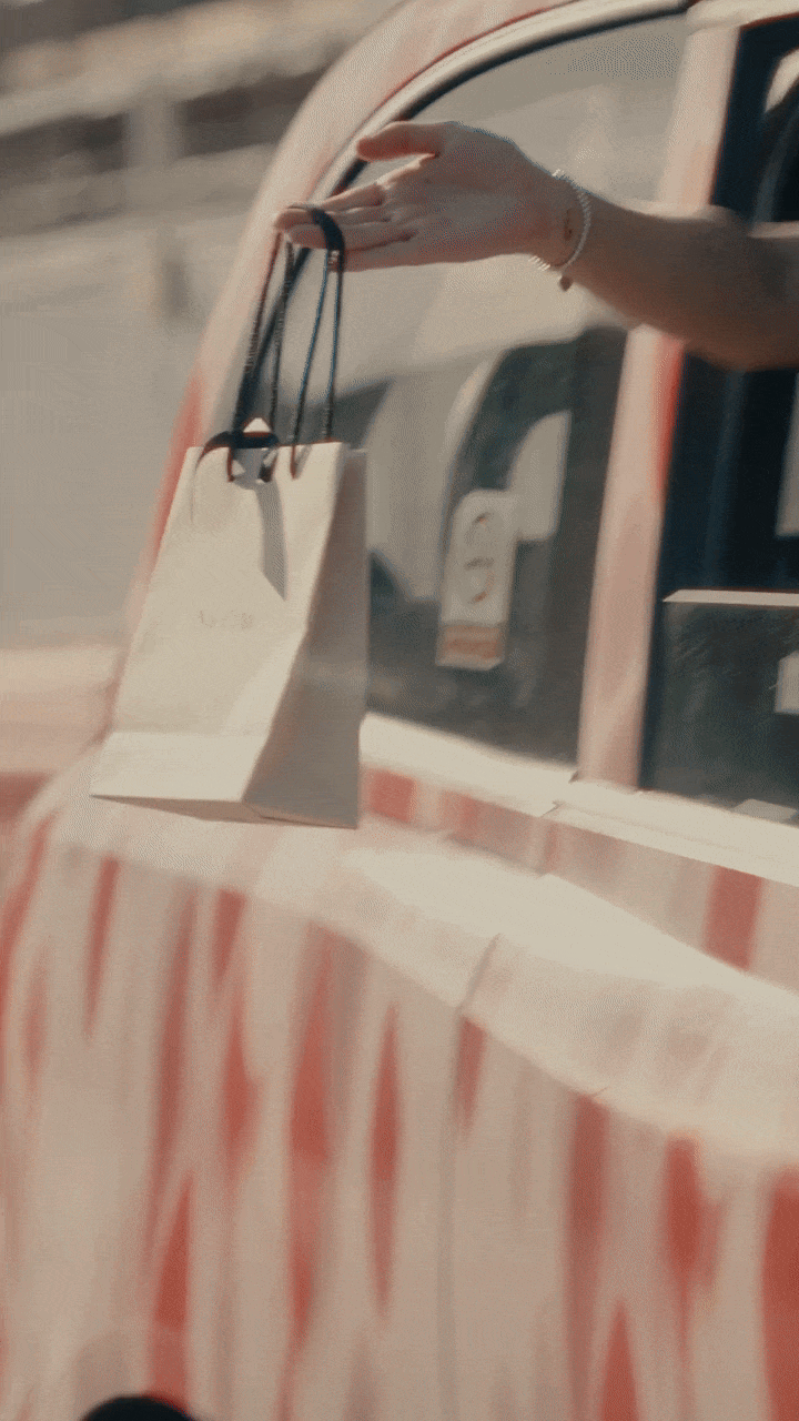 A hand extends from a vehicle window, holding a small paper bag with black string handles.