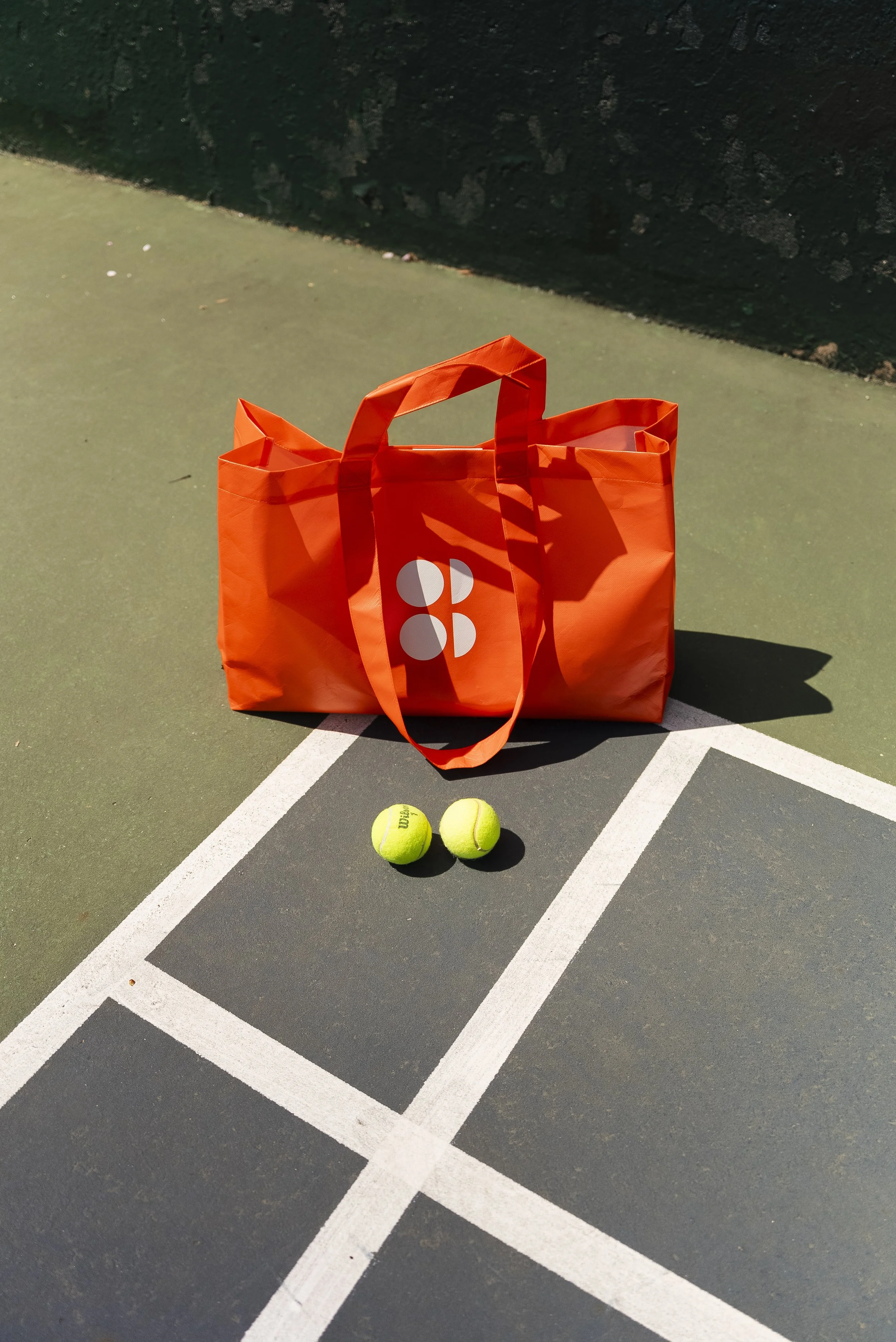 An orange sweaty betty tennis bag with a logo on it, two tennis balls, all placed on a tennis court with white lines.