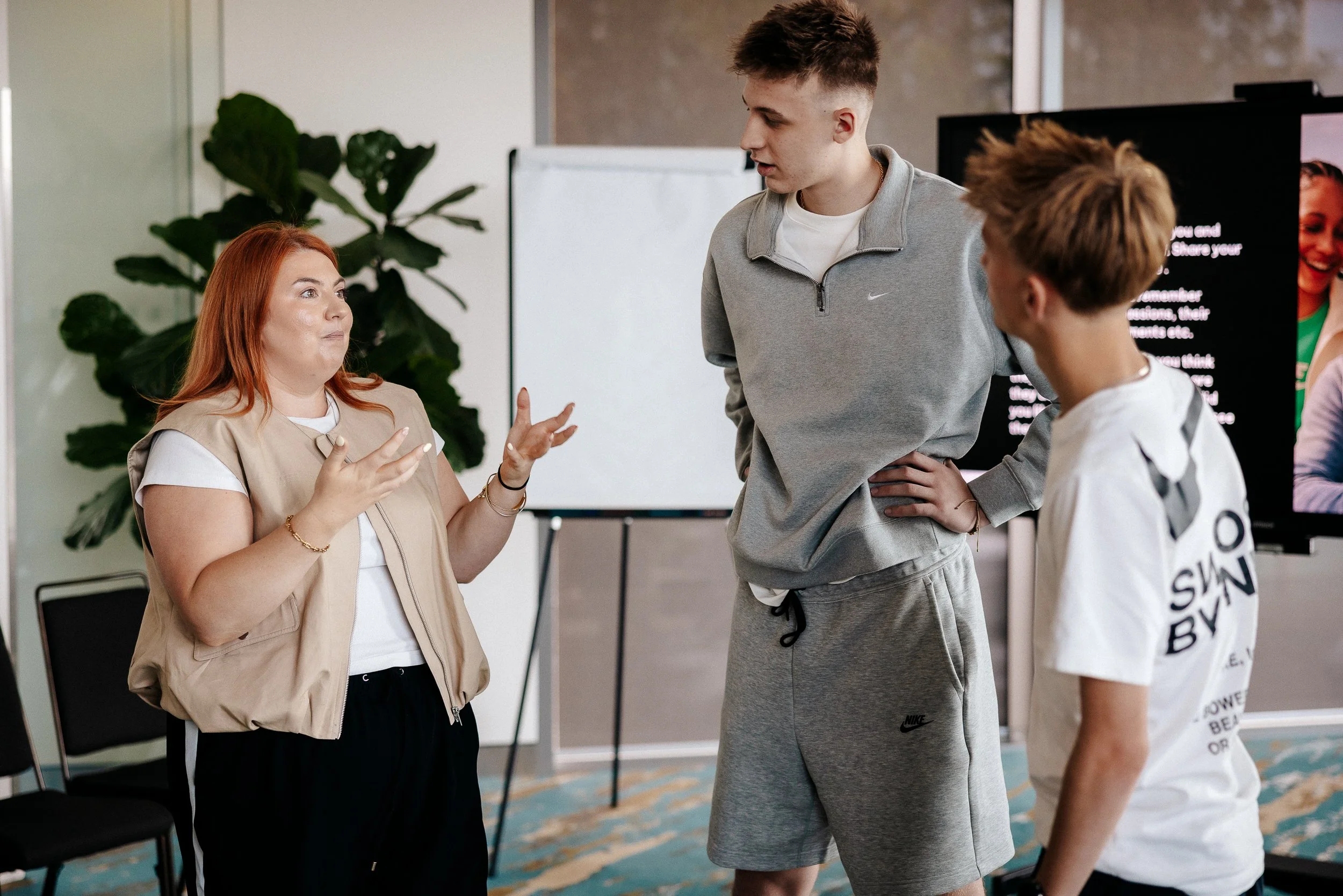 A woman with red hair is speaking to two teenage boys, one in a gray Nike hoodie and shorts, and the other in a white t-shirt with text on the back. They are in a room with a large plant, chairs, and a whiteboard, engaging in a discussion or lesson.