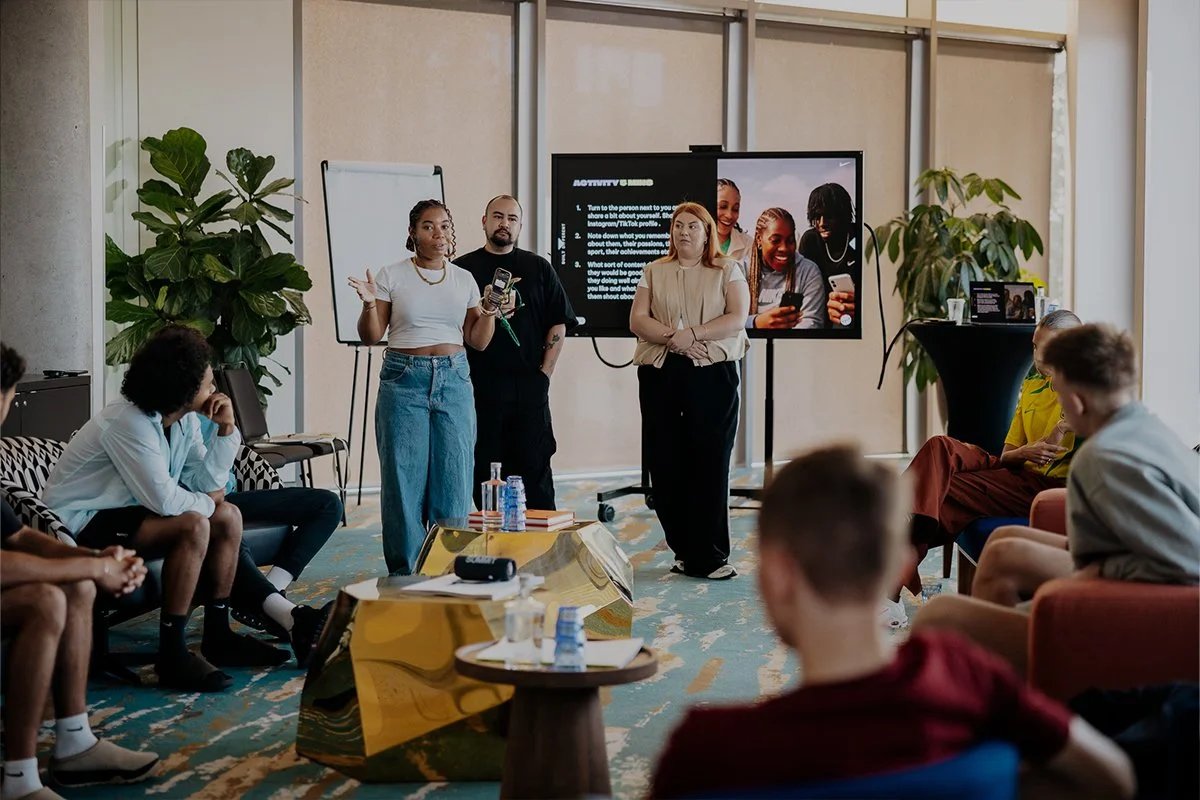 A group of people attending a presentation in a conference room, with three presenters standing at the front near a large screen displaying slides. The audience is seated and listening attentively.