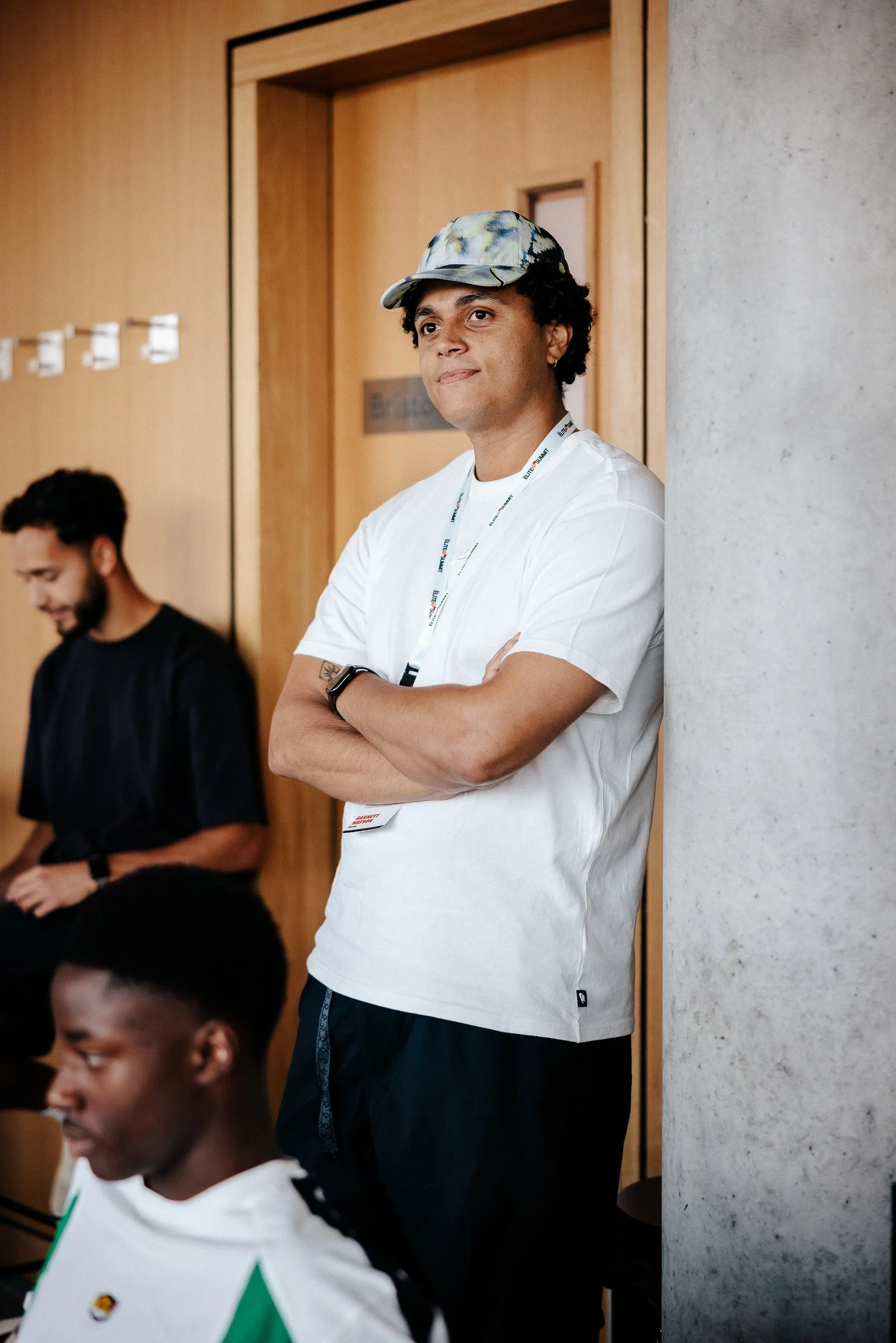 A man wearing a colorful cap and white t-shirt stands with arms crossed, leaning against a concrete support beam in a room, while two other men are seated nearby.