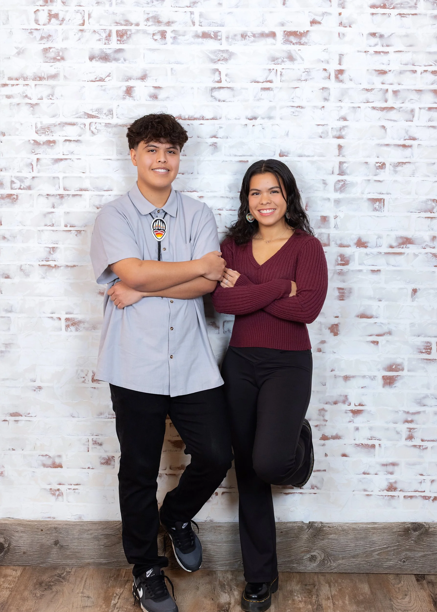 Two people posing in front of a white brick wall. The person on the left is a young man with curly dark hair, wearing a light gray button-up shirt and black pants. The person on the right is a woman with long dark hair, wearing a burgundy sweater and black pants, standing with one knee on the ground and arms crossed.
