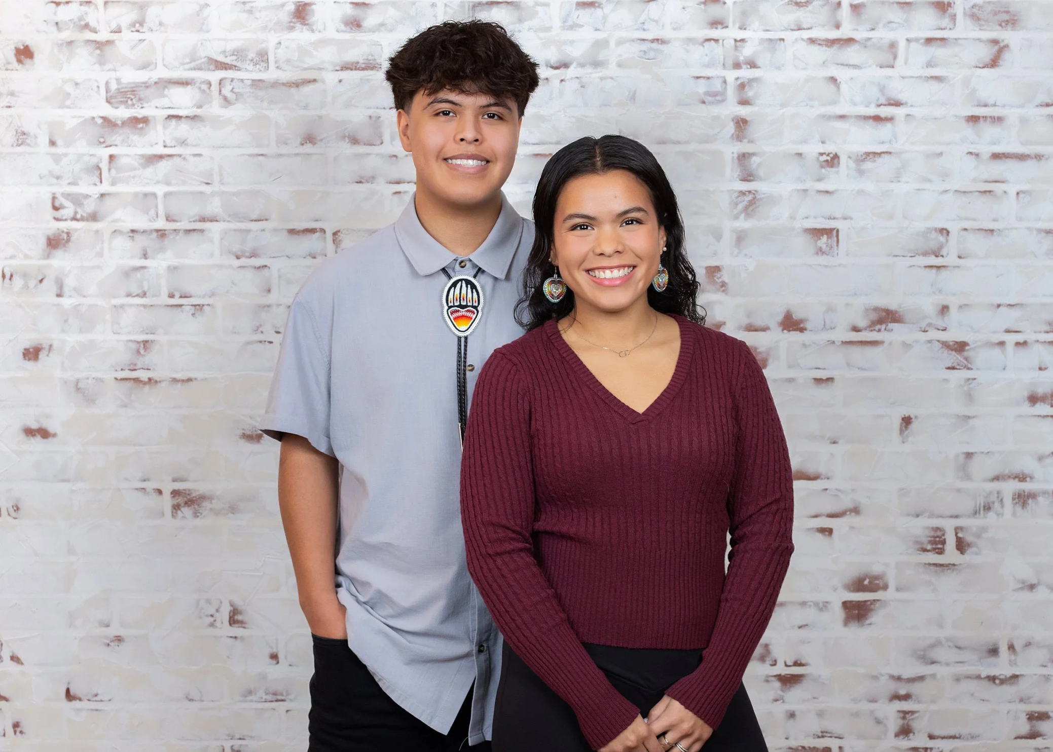 A young man in a light gray shirt with a colorful embroidered patch on the front, standing next to a young woman in a maroon V-neck sweater, both smiling in front of a white brick wall.