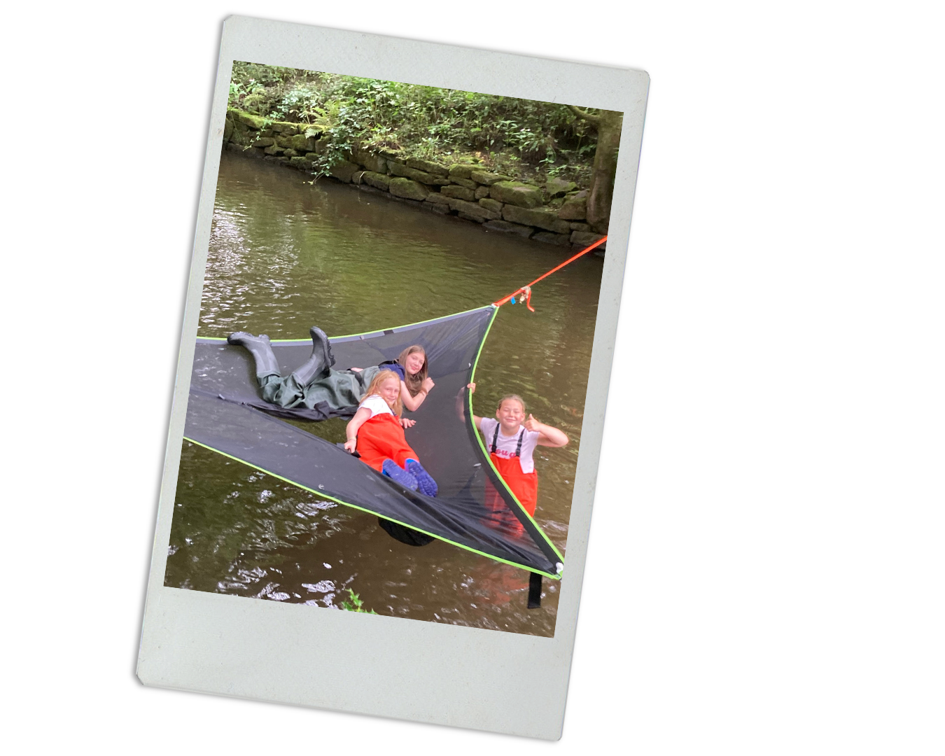 Three children are on a lake in a floating mesh tent, with one sitting on the edge, one lying down, and the third standing in the water, smiling and making a peace sign.