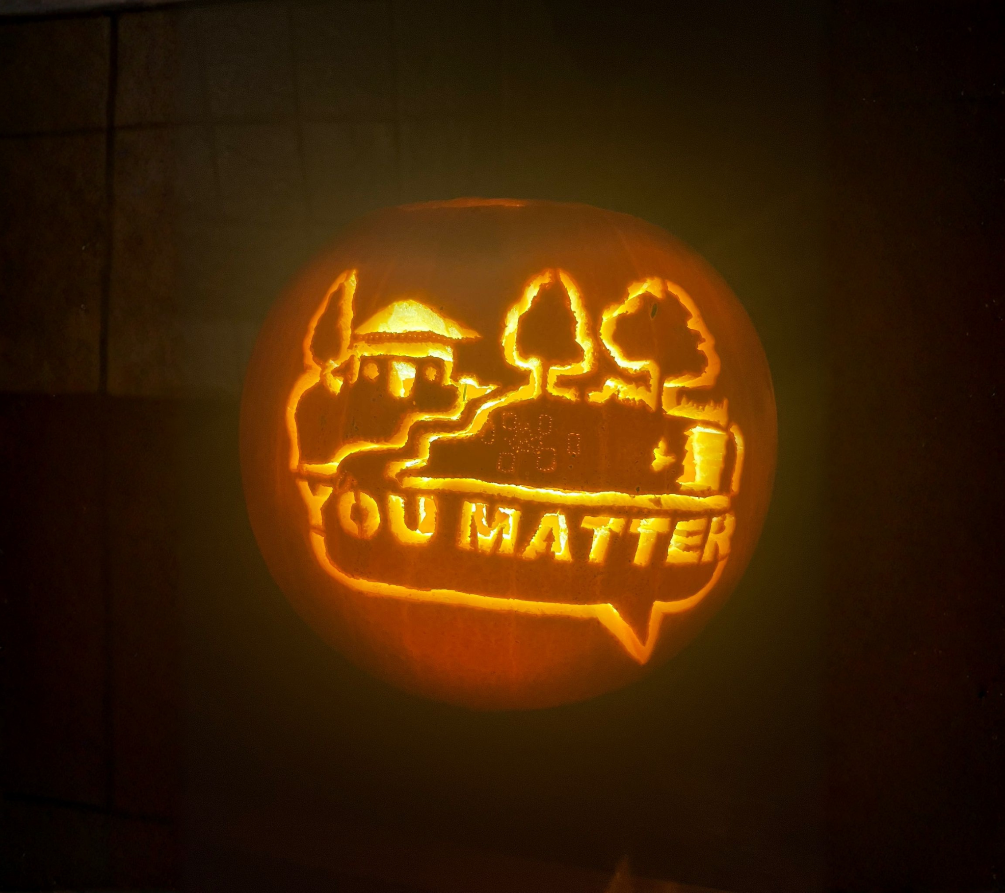 Pumpkin with a carved design of a house, trees, and the word 'OUR MATT' lit from inside.