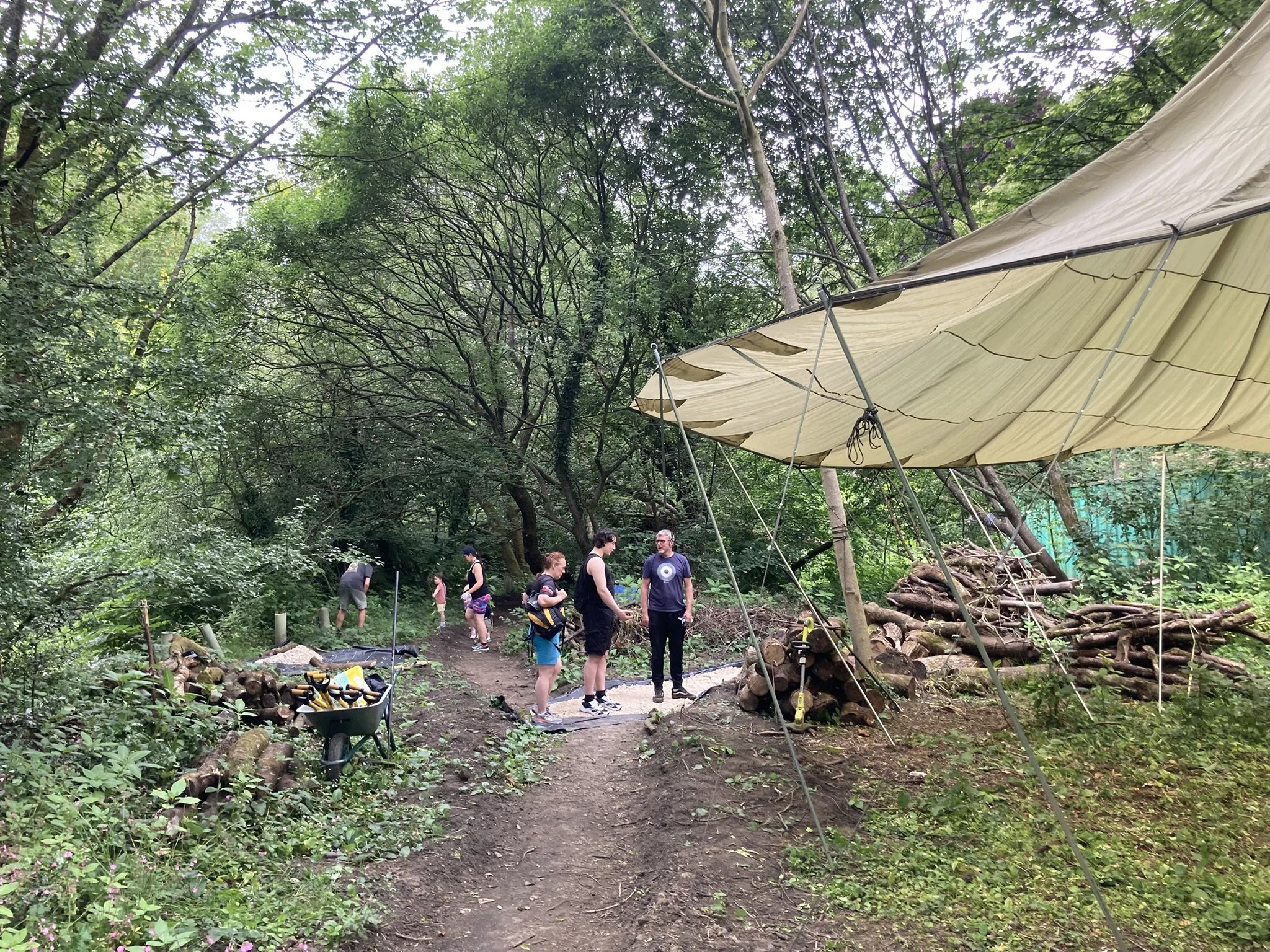 People building a large tent in a forested outdoor area with green trees, logs, and tools nearby.