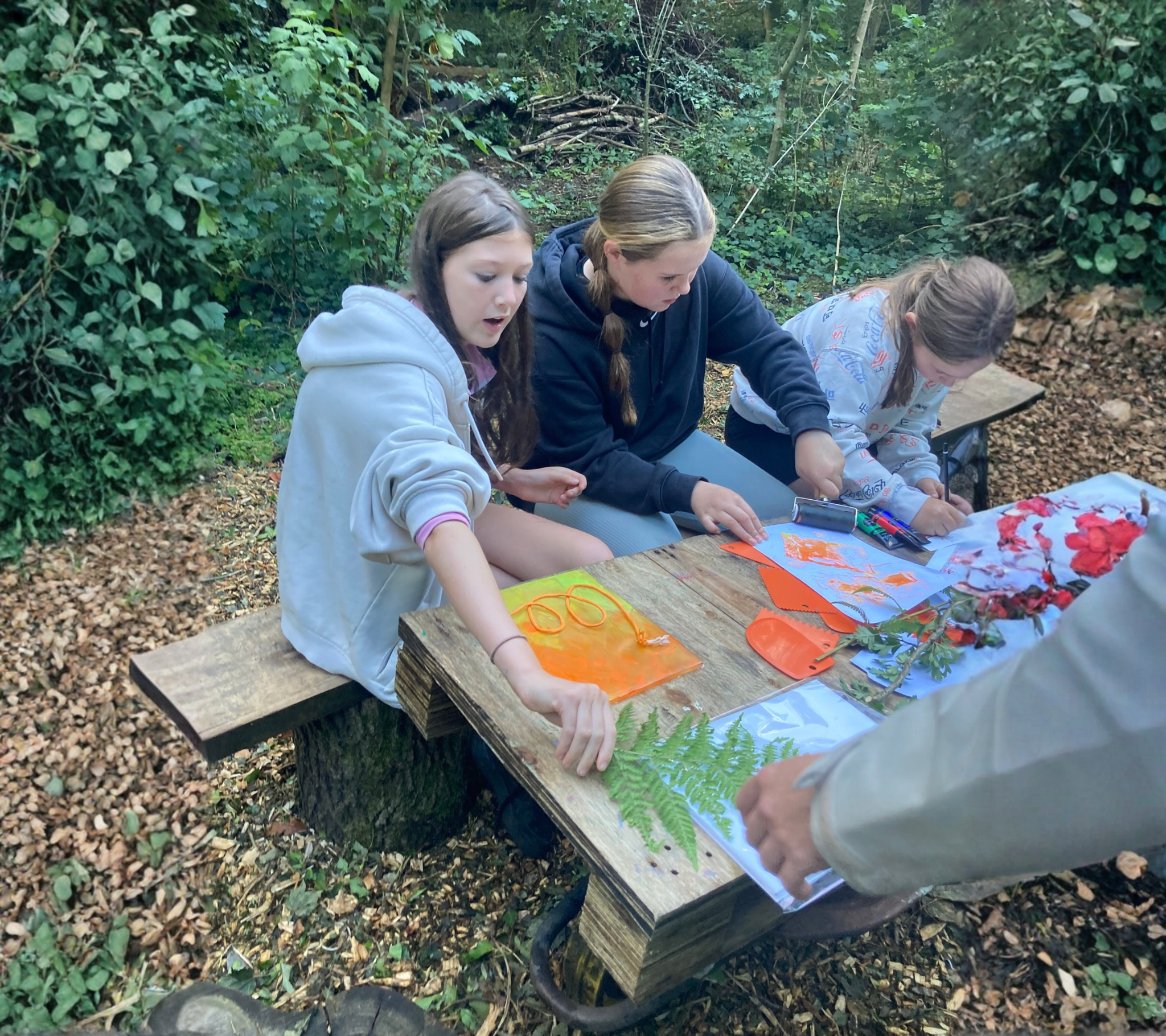 Three young girls sitting at a wooden table outdoors, engaged in nature craft activities with leaves, flowers, and markers surrounded by dense green foliage.