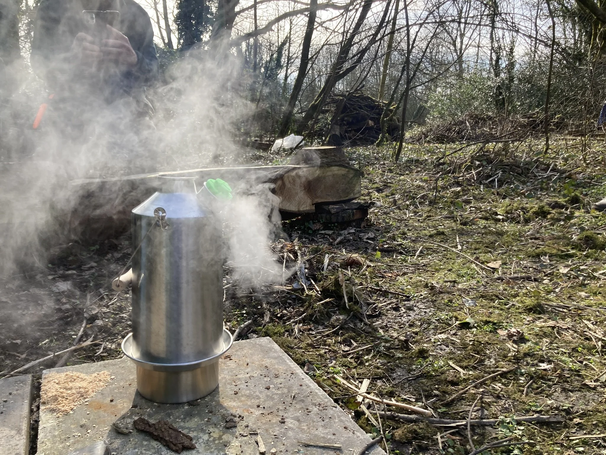 A camping cooking kettle on a table outdoors with steam rising from it, surrounded by a wooded area with fallen branches and moss on the ground.