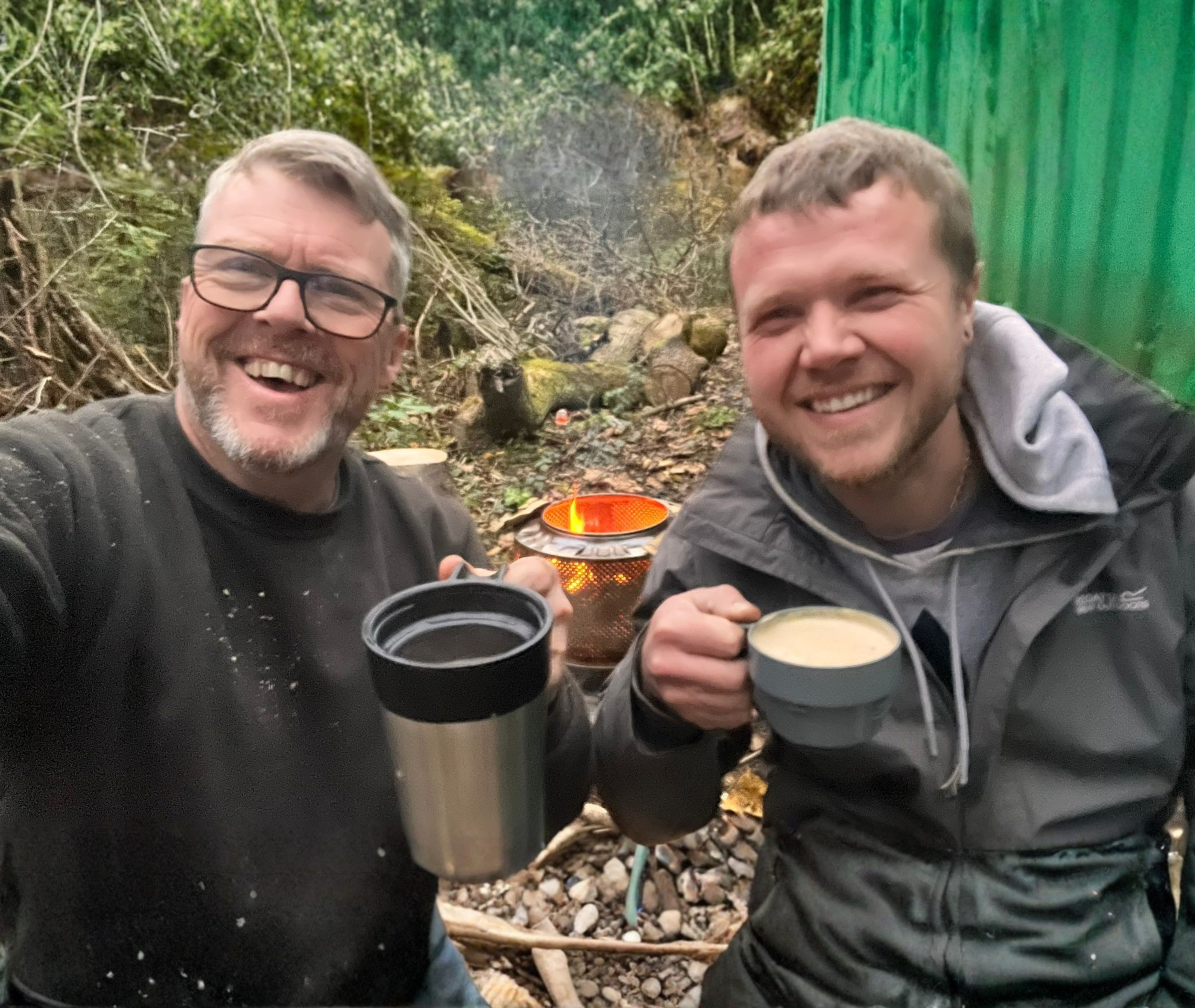 Two men smiling outdoors, holding cups of coffee, with a campfire and green fence in the background.