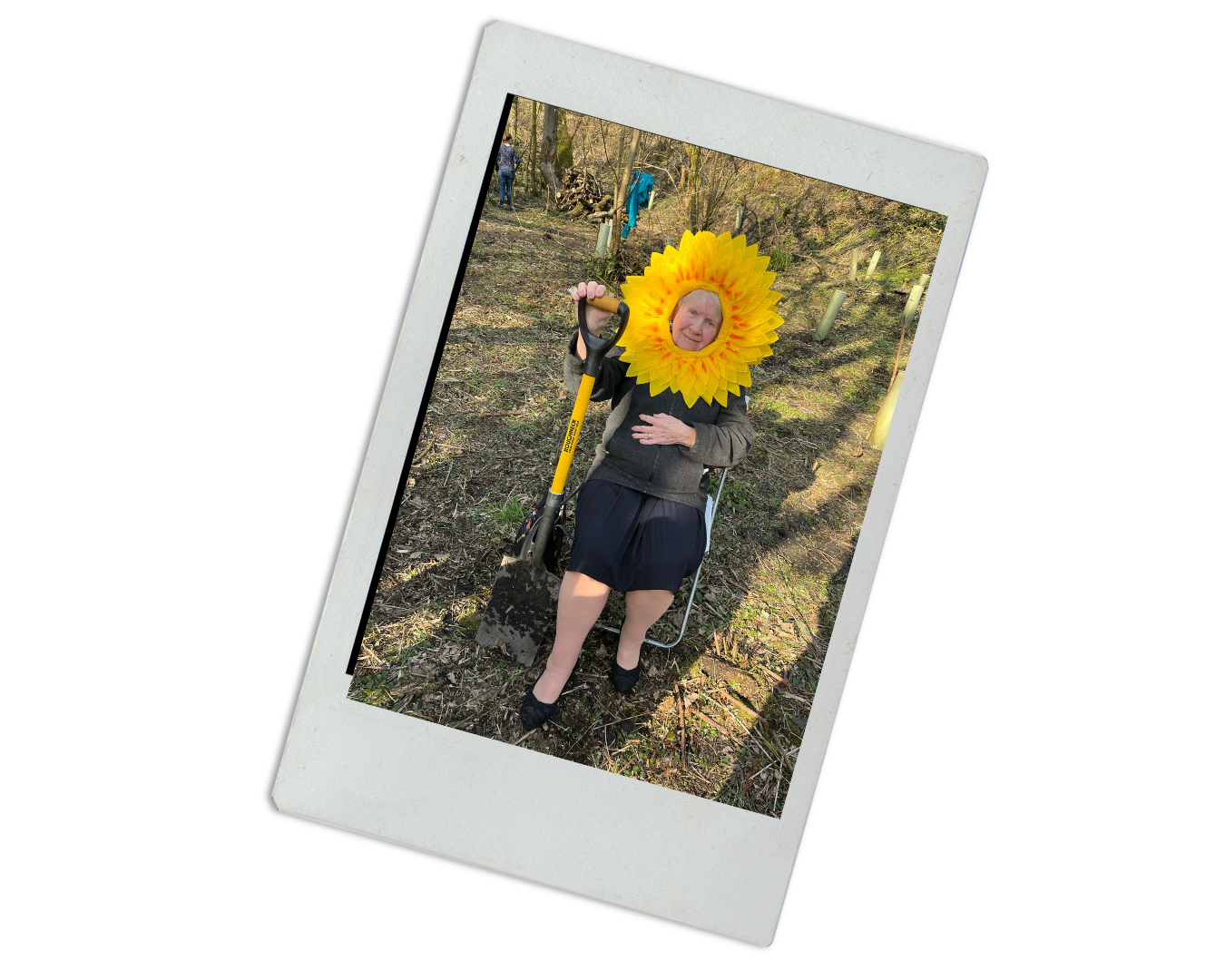 Person wearing a sunflower costume in a garden or field, holding a shovel, with two other people in the background.