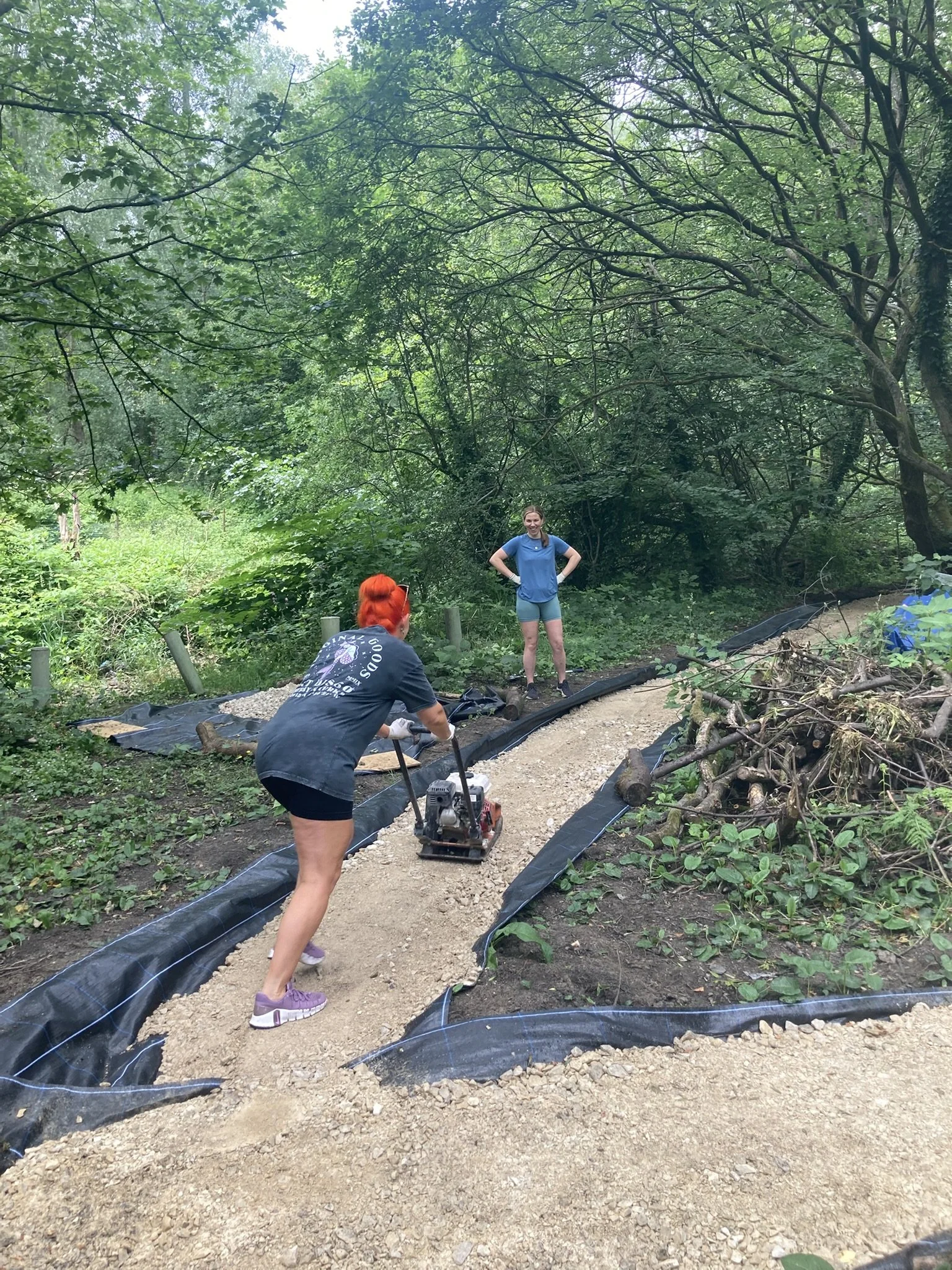 Two women working on a trail in a lush green forest, one using a compactor, the other standing with hands on hips.