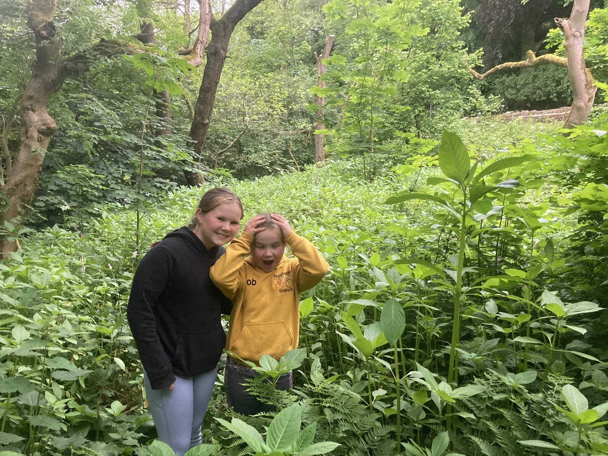 Two young girls smiling and reacting with surprise in a lush, green forest with tall trees and dense plants.