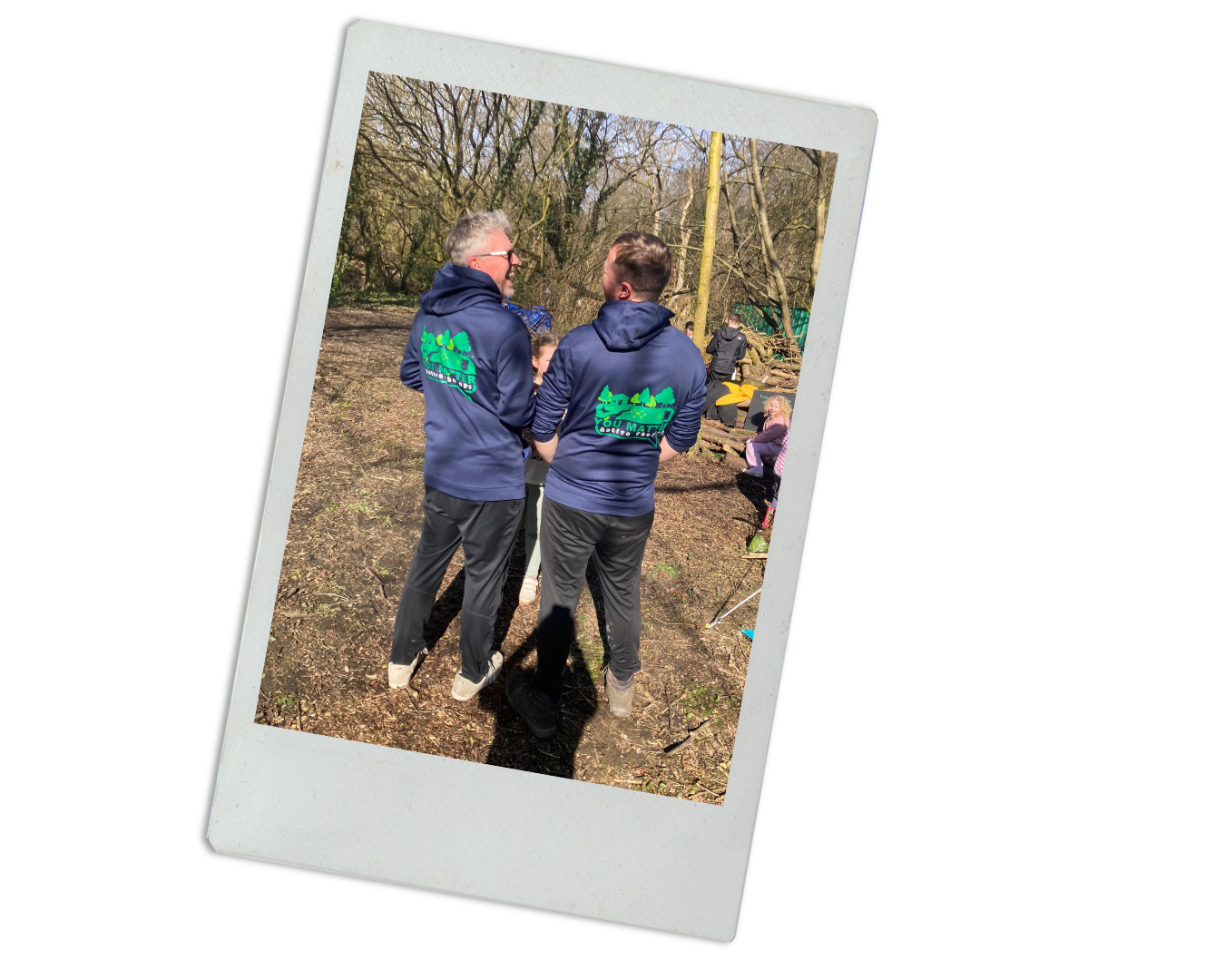 Two men wearing navy blue hoodies with green logo and text on the back, standing outdoors in a wooded area, engaged in conversation. Several other people are in the background, seated on logs or standing among trees.