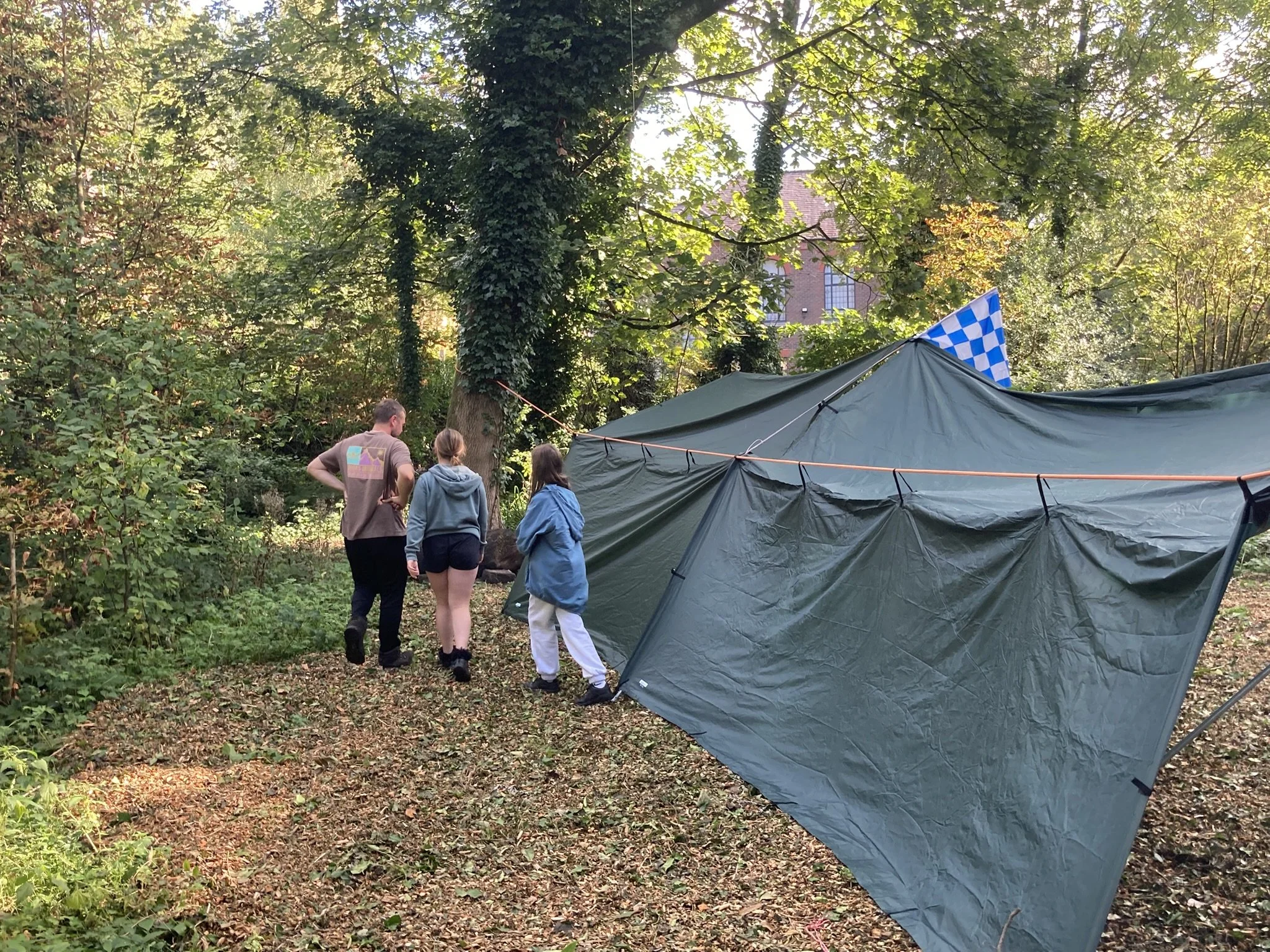 Three people walking through a wooded area near a large green camping tent with a blue and white checkered flag.