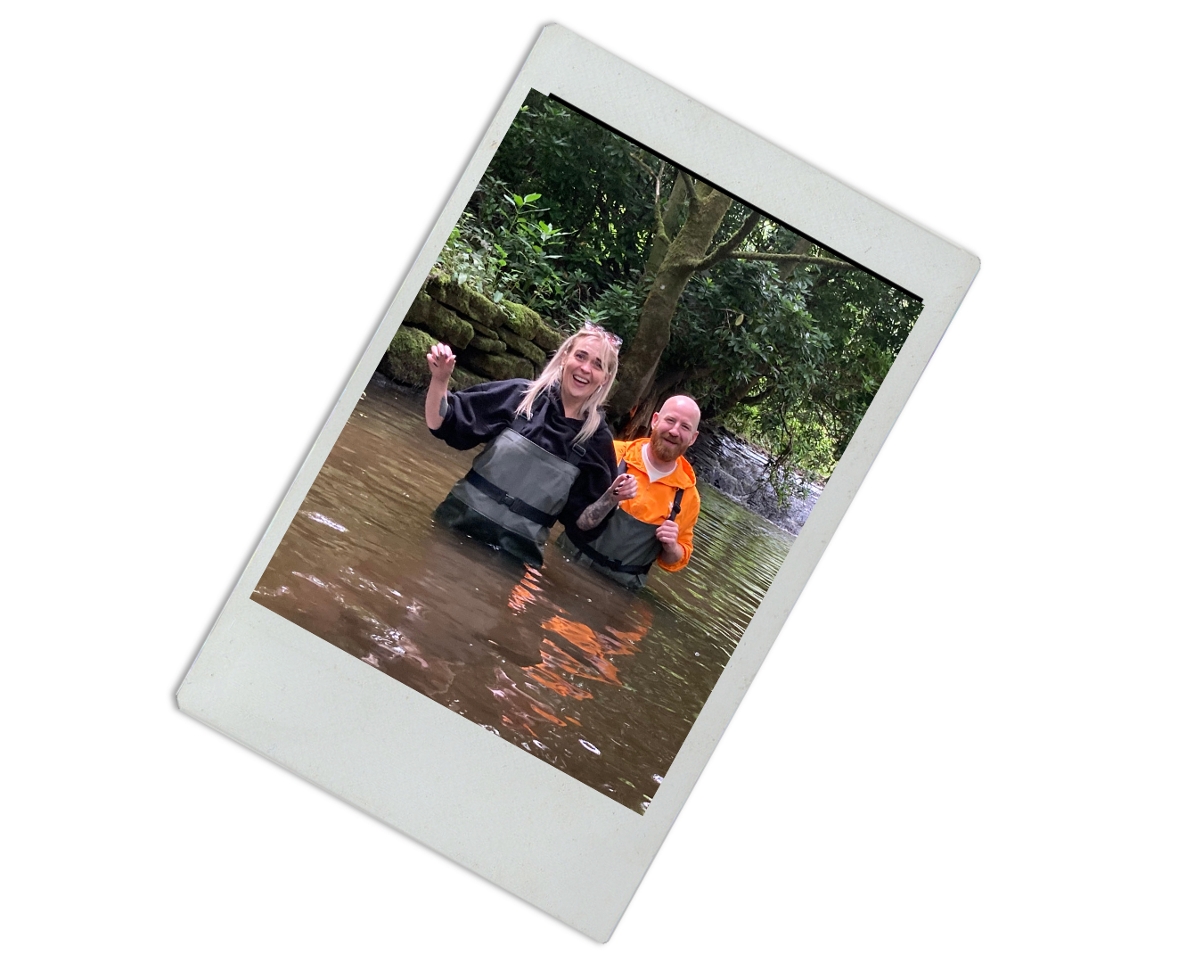 Two smiling people standing in a river, holding their backpacks, with trees and rocks in the background.