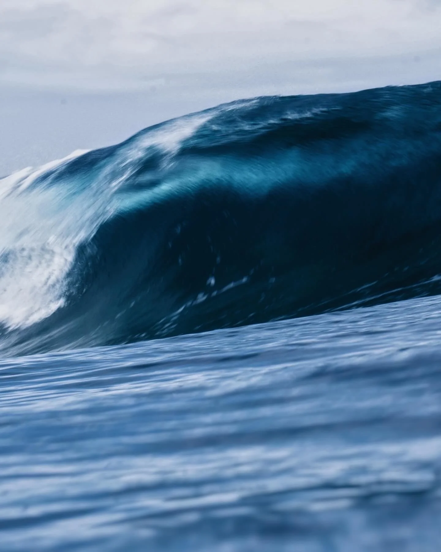 A large ocean wave with white foam on the crest, viewed in the open sea with a cloudy sky in the background.
