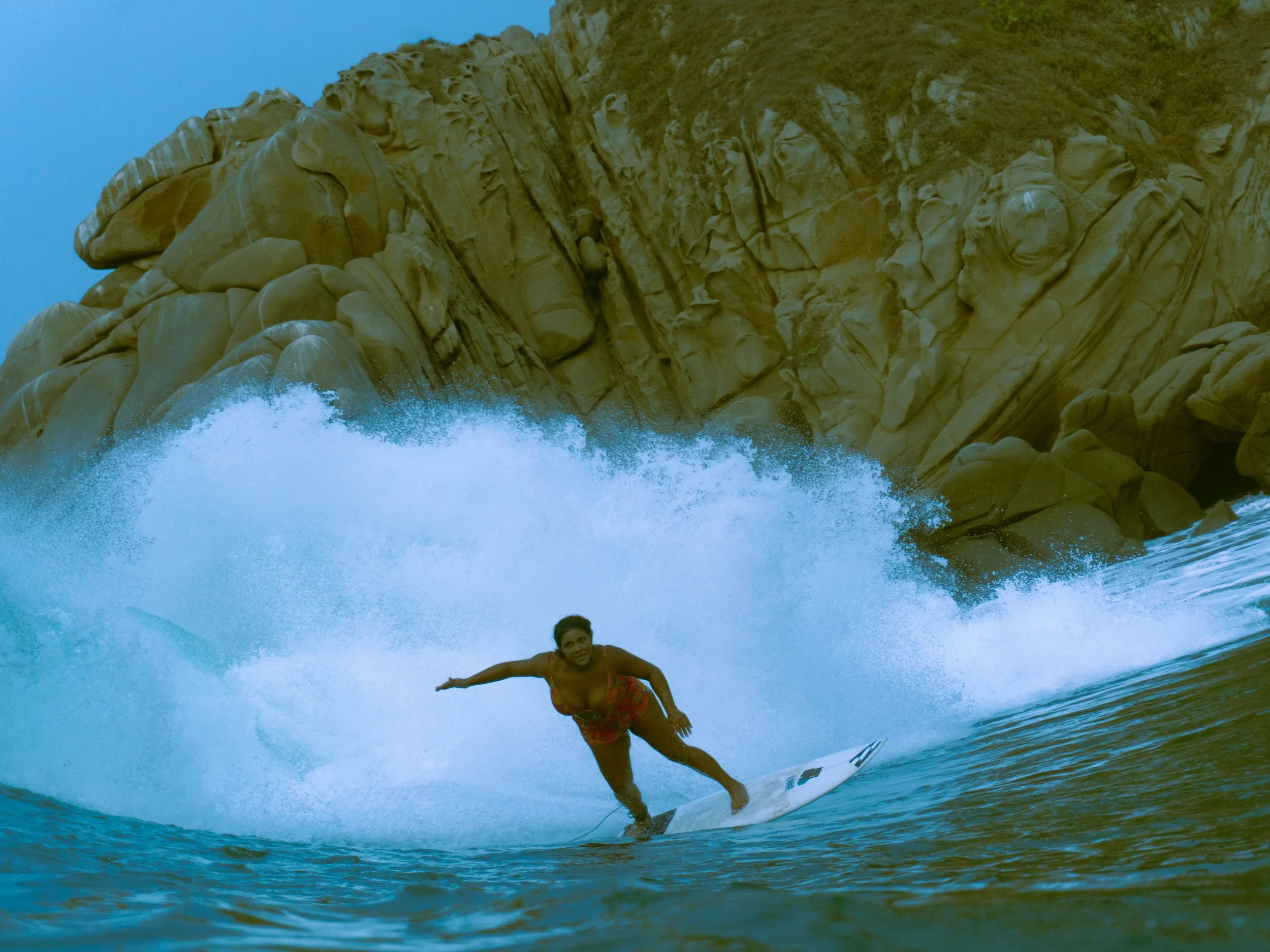 A woman surfing on a wave near rocky shoreline with large boulders.