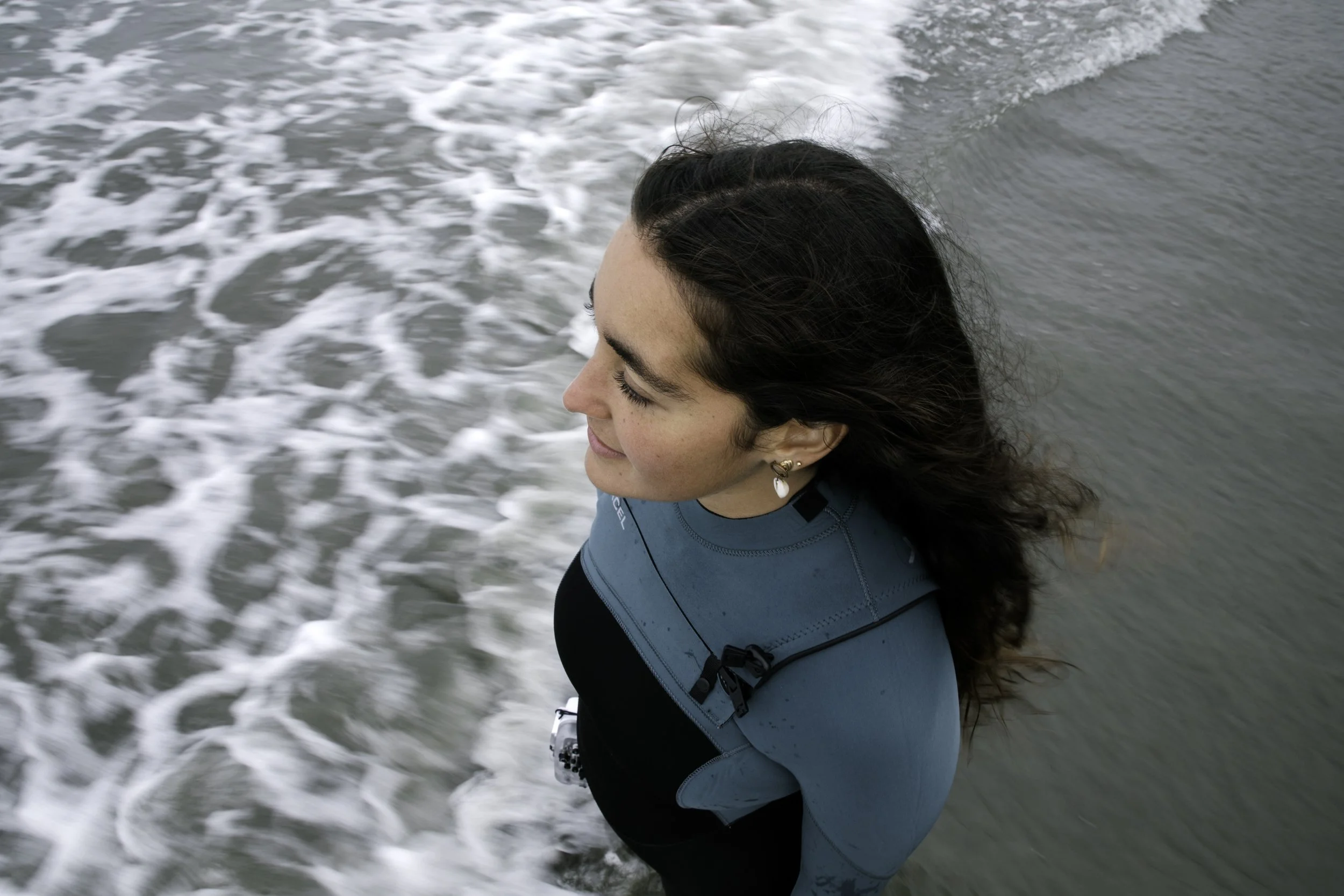 A woman with dark, wavy hair and earrings standing on the beach with ocean waves behind her, smiling with eyes closed.