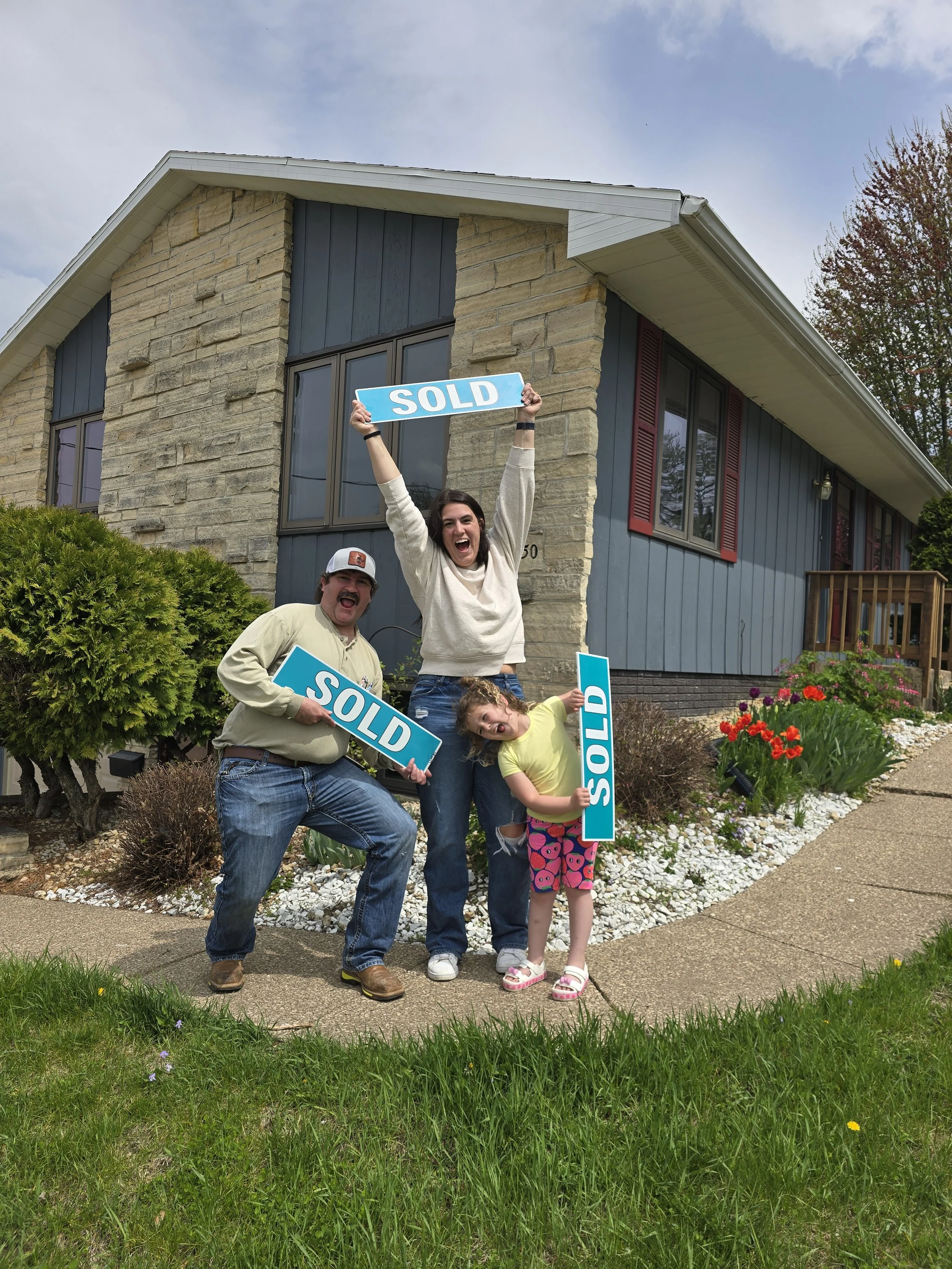 A family of three celebrating in front of a house with 'SOLD' signs, indicating they bought the home.