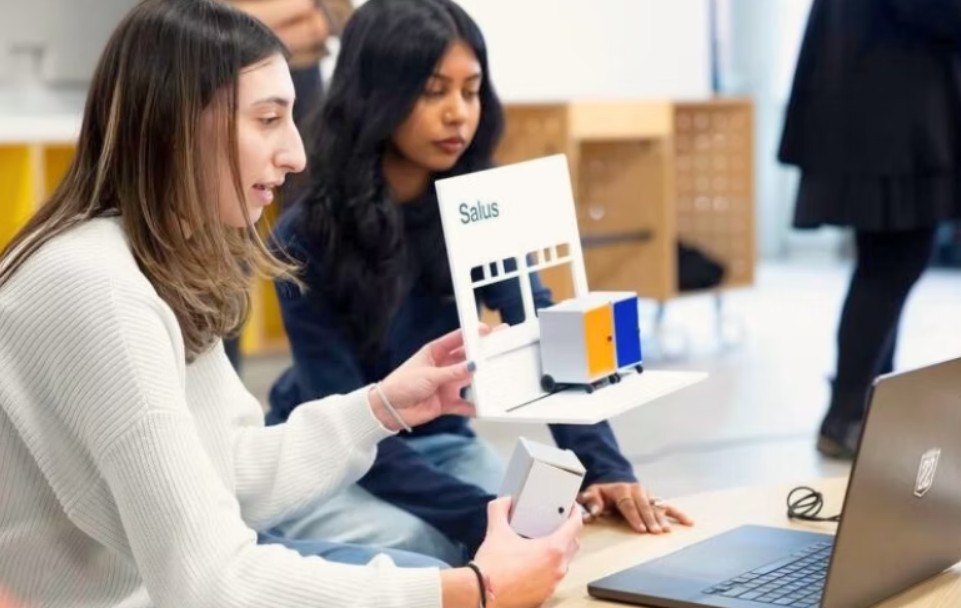 Two women sitting at a table, one holding a model of a Salus-branded device or structure, with a laptop nearby, in an indoor setting.