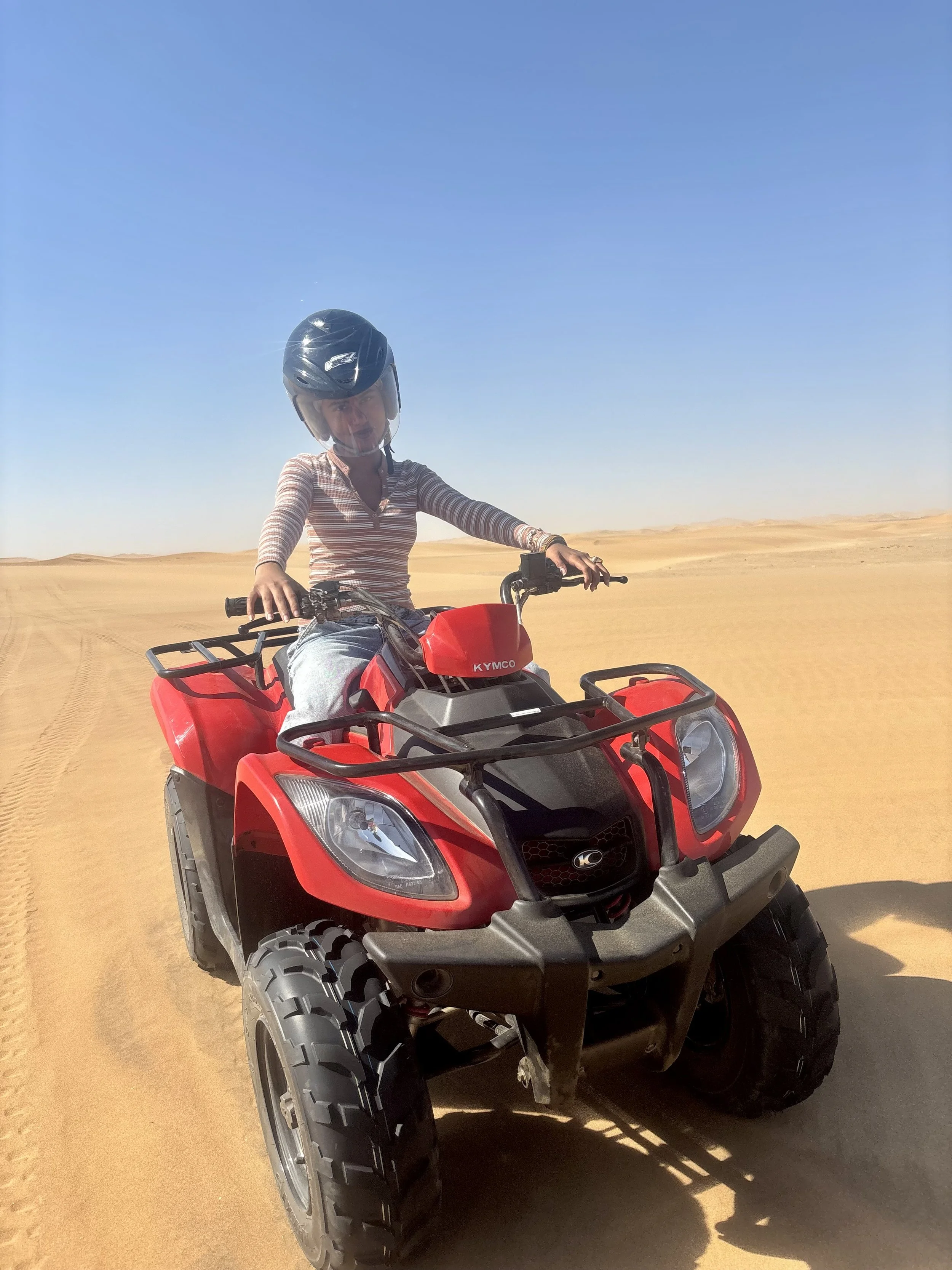 Person wearing a helmet riding a red ATV across a desert landscape with sand dunes and a clear blue sky.