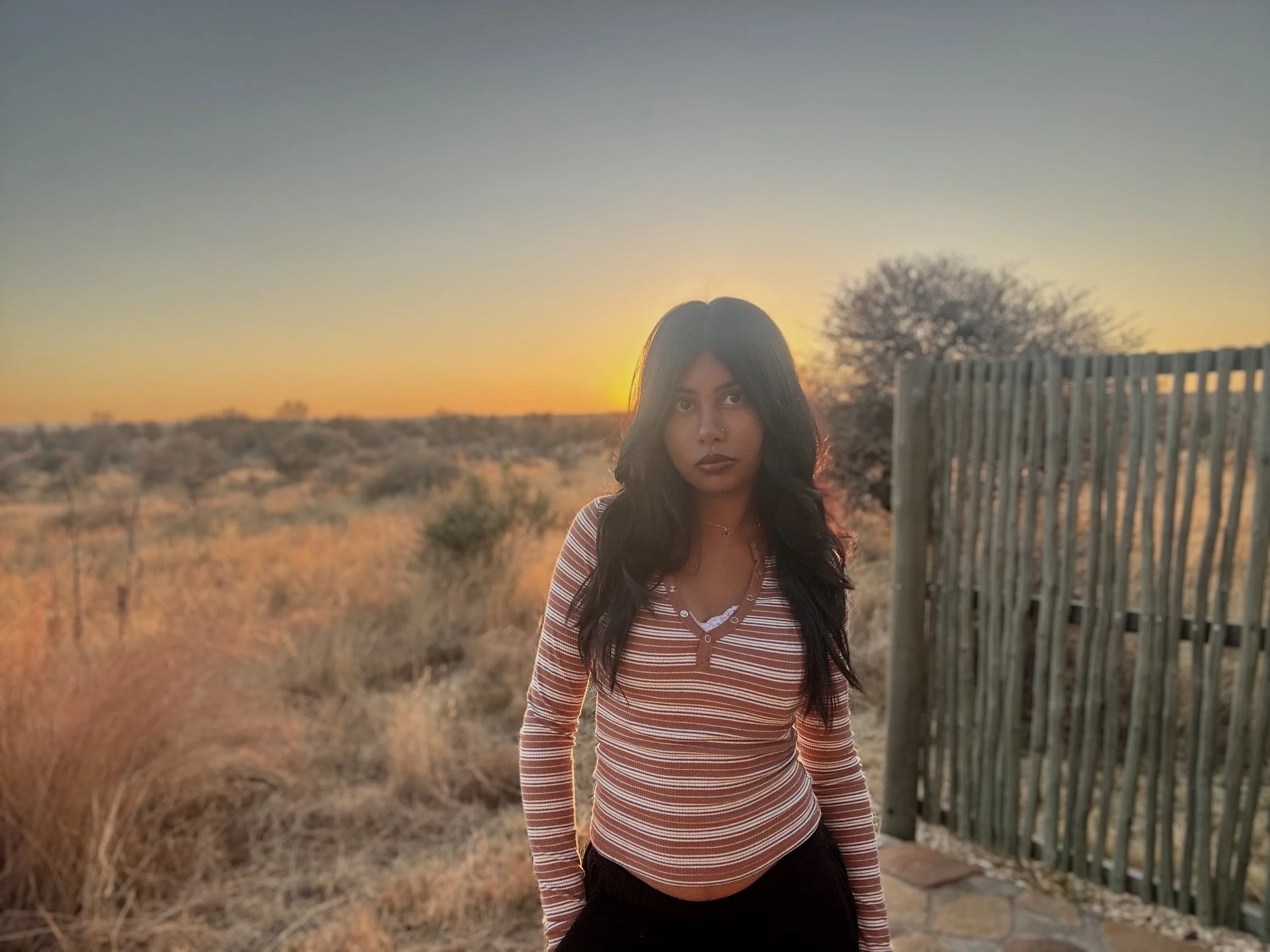 Young woman with long dark hair standing outdoors during sunset in a dry, desert-like landscape with bushes and a fence nearby