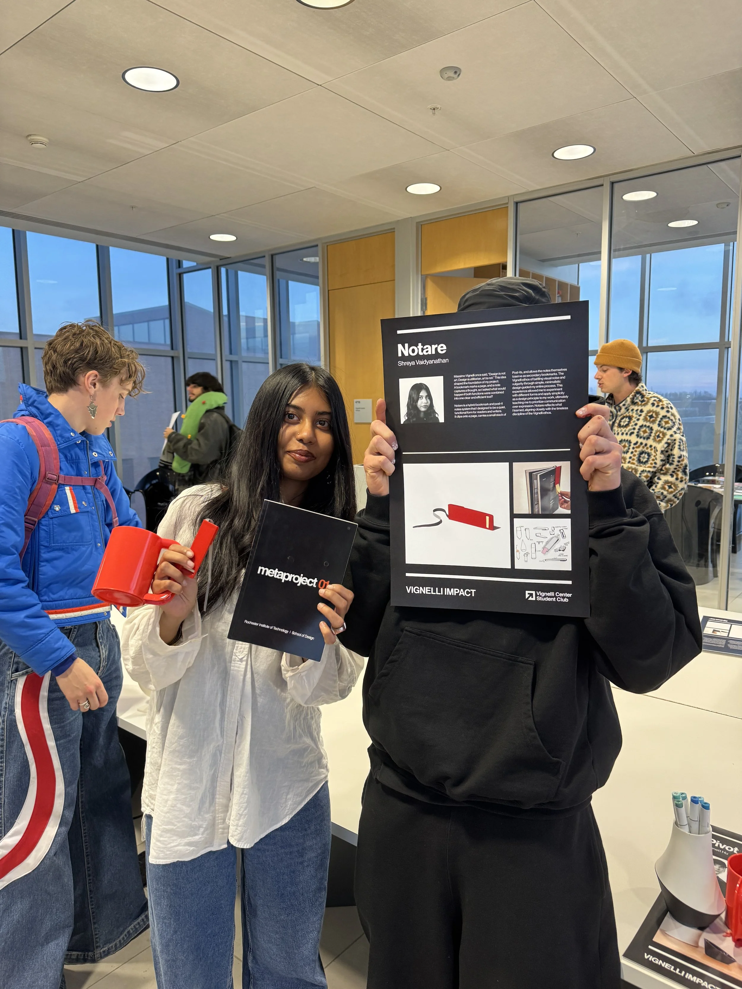 Two women stand indoors, one holding a black booklet titled 'metaproject' and the other holding a large display board with information and images about a person named 'Notare'. Several other people are visible in the background, with large windows and modern decor.