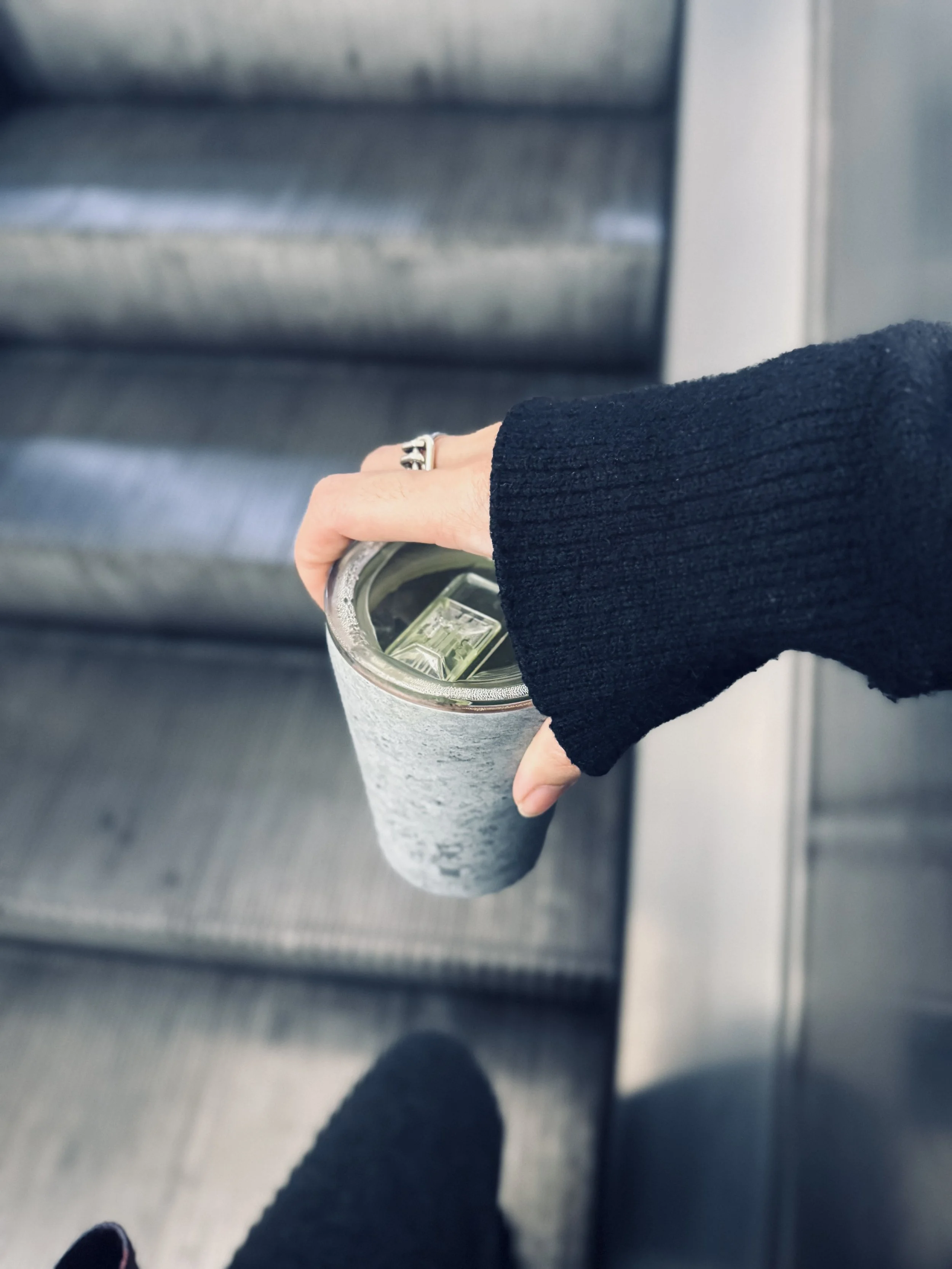 A person's hand holding a large insulated tumbler with an open lid, containing a cold beverage with ice cubes, in an indoor setting.