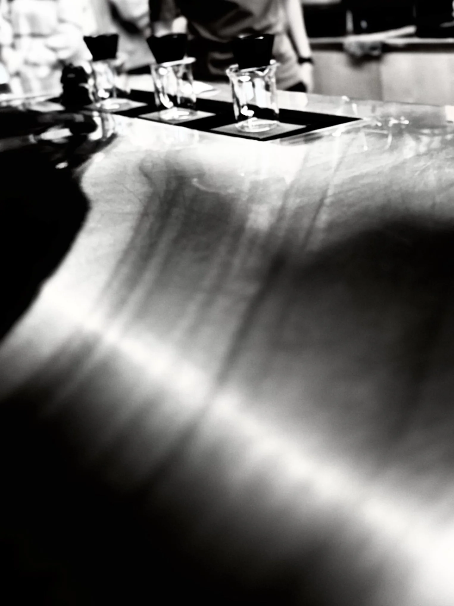 A black and white photo showing a row of shot glasses on a table, with some people in the background.