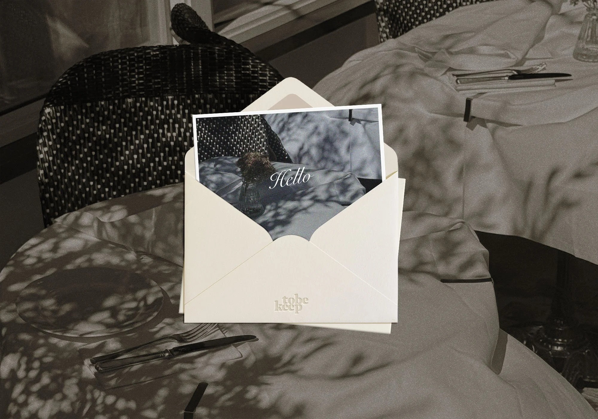 A white greeting card in an open envelope with the word 'Hello' on it, placed on a table next to cutlery and a plate, with shadows of leaves creating a pattern on the tablecloth.