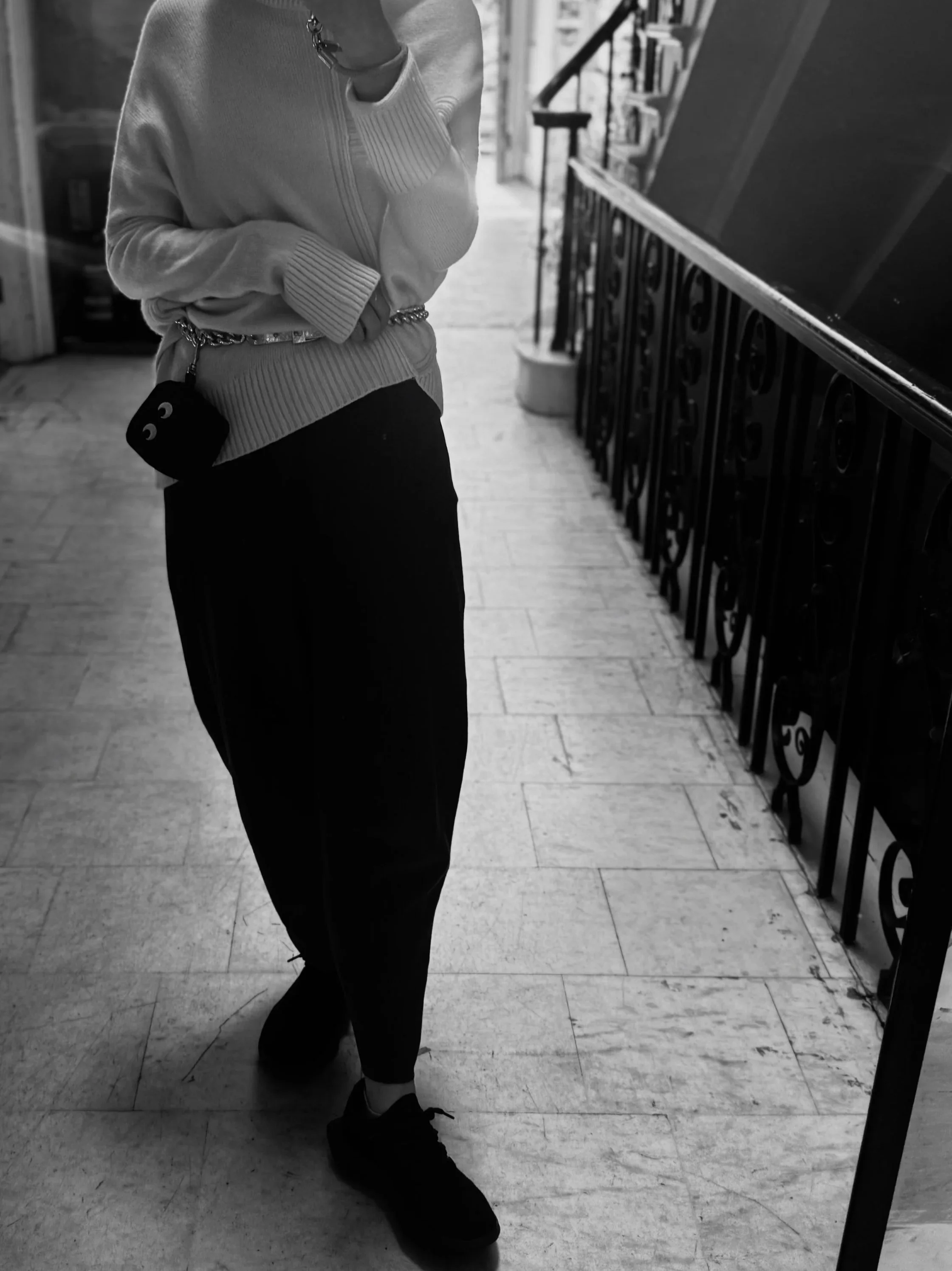 A person taking a mirror selfie, wearing a light-colored sweater, dark pants, sneakers, and a small bag with a face design. The setting appears to be indoors with a staircase in the background.