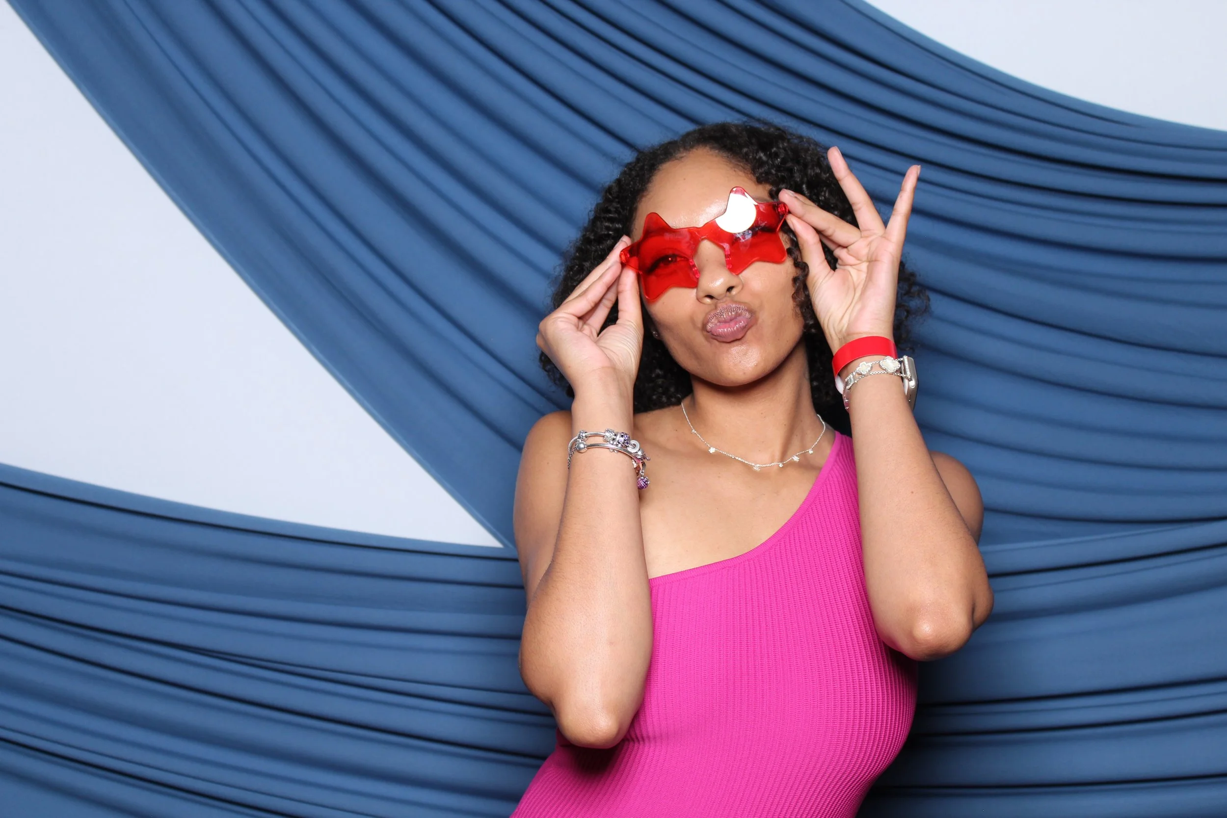A woman wearing red star-shaped sunglasses as a fin photobooth prop.  She is holding the sunglasses with both hands and making a kissy face. She stands against a beautiful draped blue backdrop.