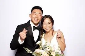 A happy couple dressed in wedding attire, smiling and holding flowers, posing against a plain white background.