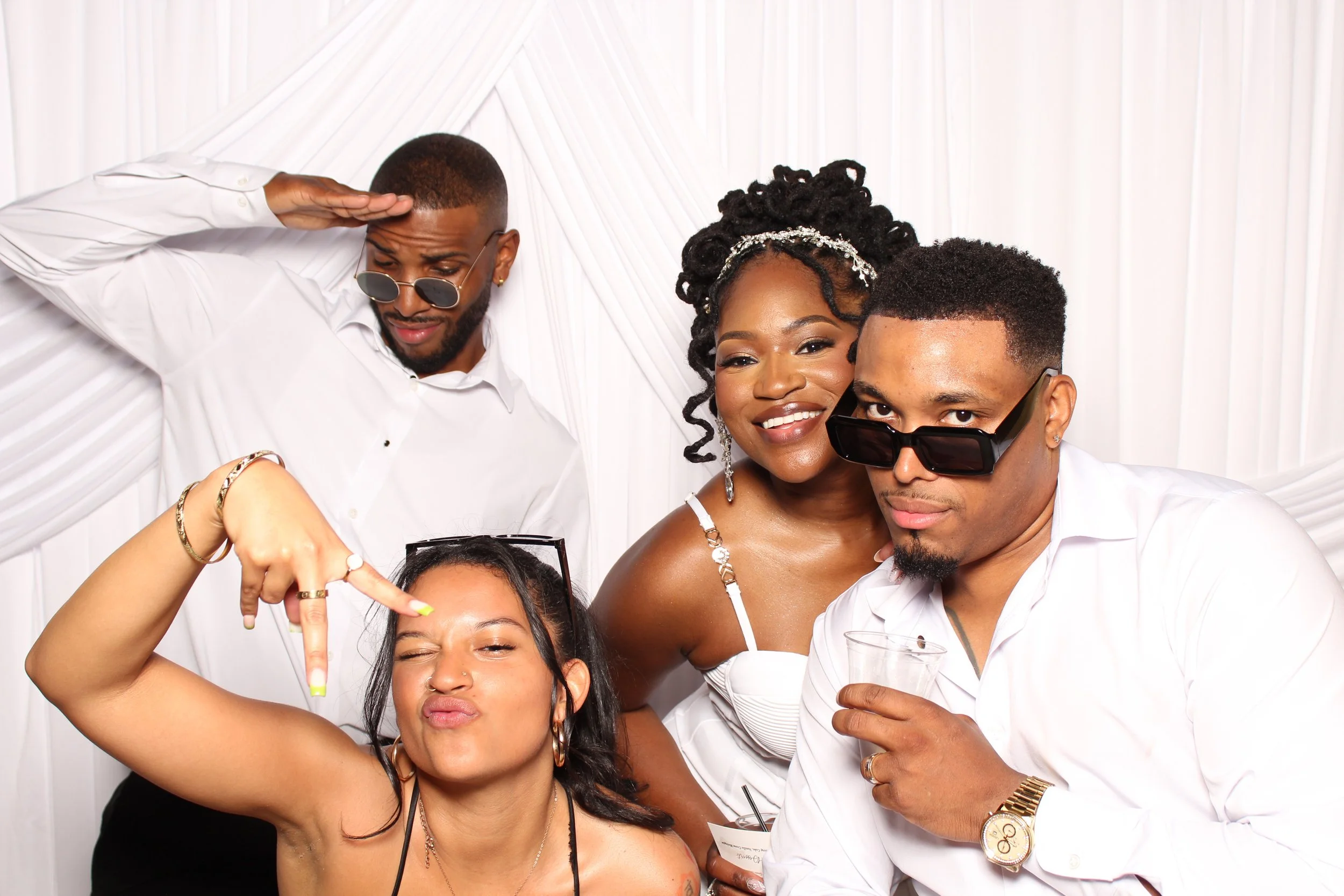 Group of five young adults taking a selfie together, some wearing sunglasses, smiling, and having fun in front of a white backdrop.