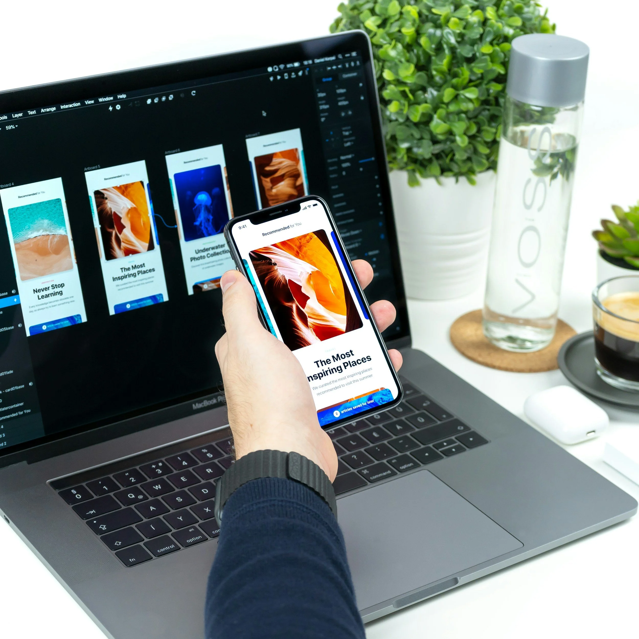 A person holds a smartphone displaying a travel app with the title 'The Most Inspiring Places.' The phone is held above a laptop showing the same app design. On the desk, there is a water bottle, a potted plant, a coffee drink, and a white wireless earbud case.