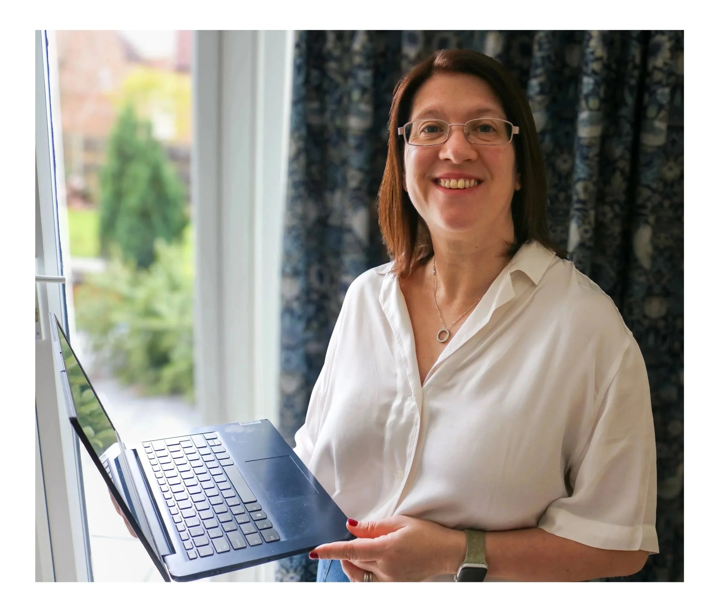 Digital Goat Founder Sarah with shoulder-length brown hair and glasses smiling while holding an open laptop near a window with greenery outside is introducing you to her digital marketing website for website auditing and website creation