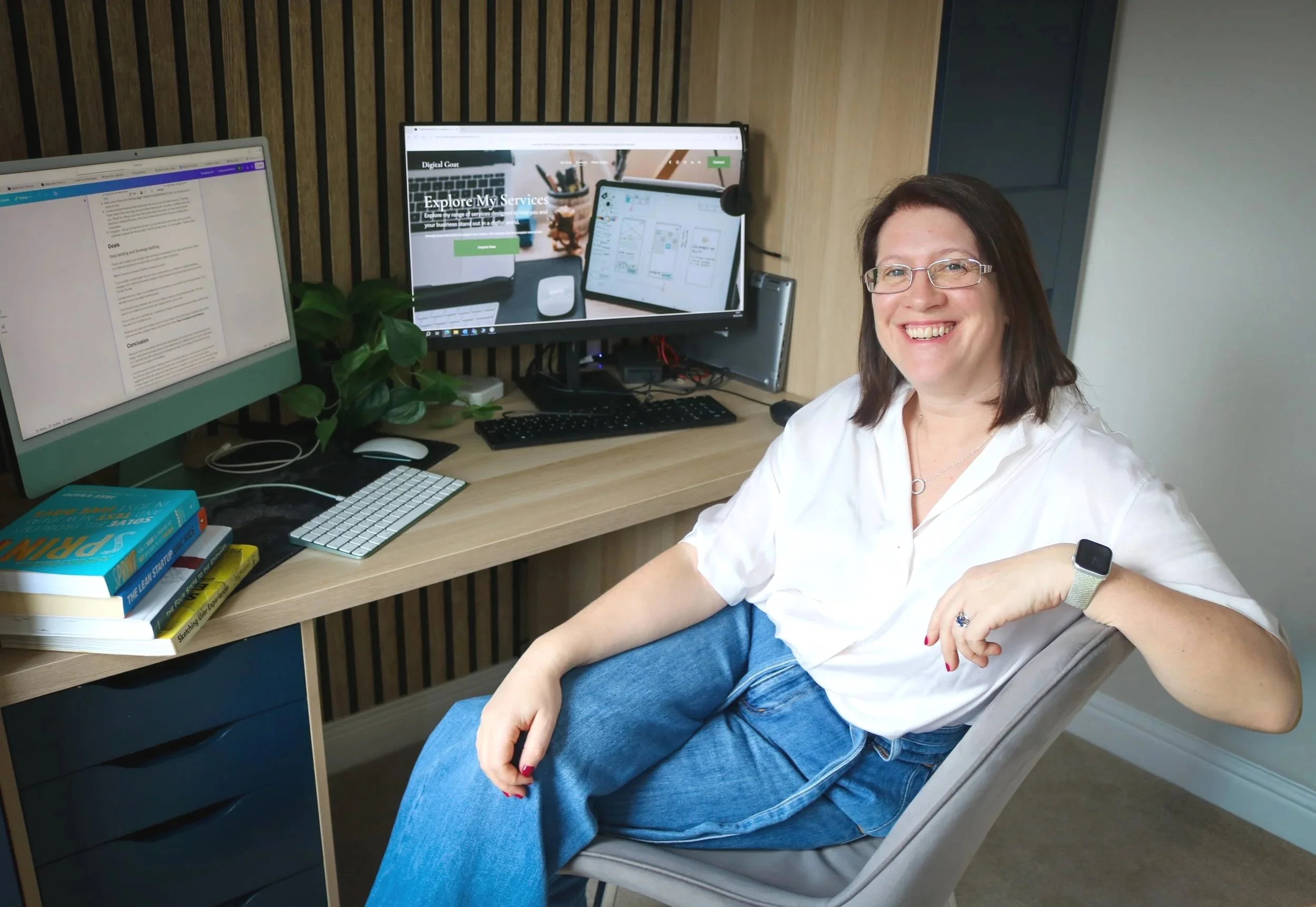 Digital Goat Founder Sarah with glasses and a white shirt sitting at a desk with dual monitors, surrounded by books, a keyboard, and a potted plant, smiling at the camera. She is introducing you to her digital marketing website home page