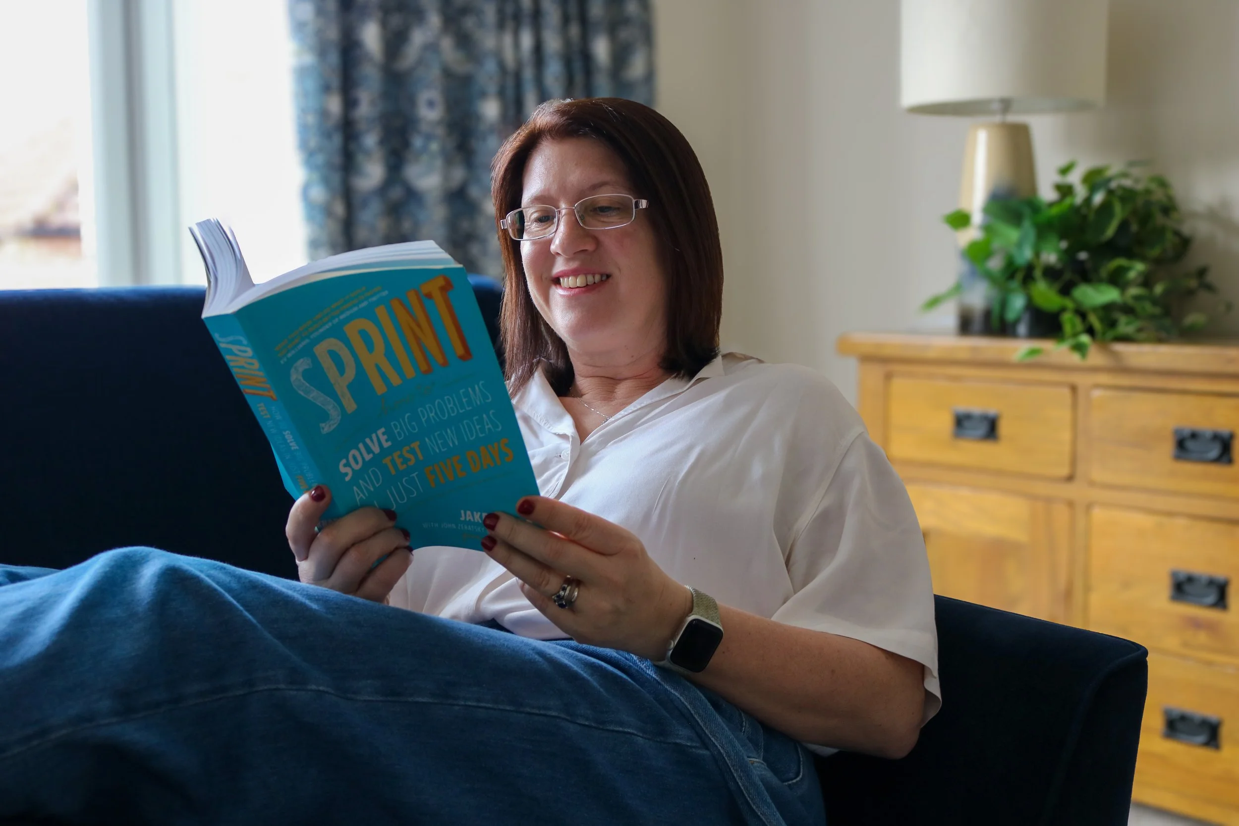 Digital Goat Founder Sarah wearing a white shirt, sitting on couch, reading a colorful book titled 'SPRINT'. She is smiling and appears relaxed. She is researching User Experiences and User Experience Design