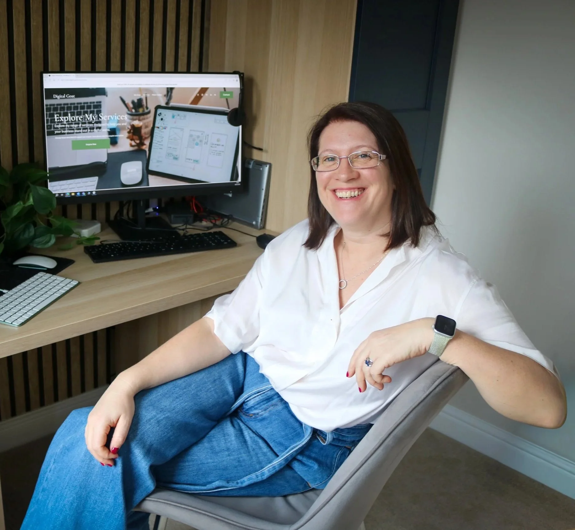Digital Goat Founder Sarah with glasses and a white shirt sitting at a desk with dual monitors, surrounded by books, a keyboard, and a potted plant, smiling at the camera. She is introducing you to her digital marketing website home page