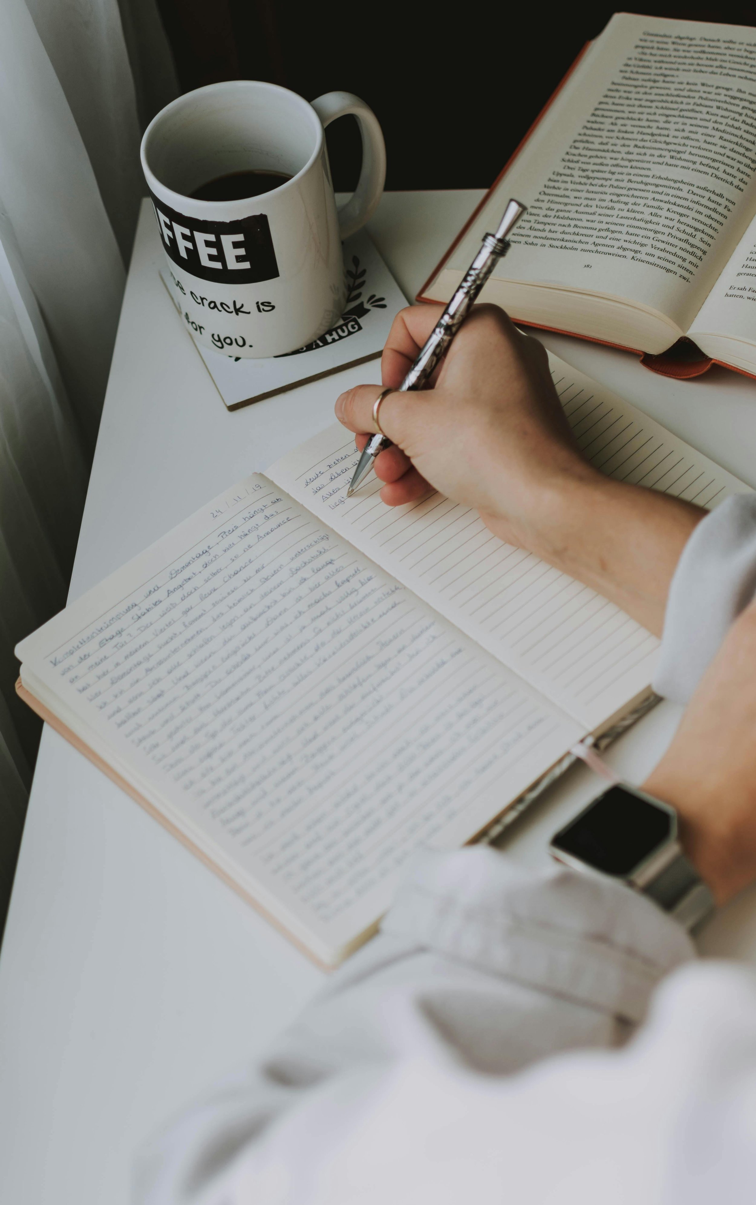 Person writing in a notebook at a white desk, with an open book, a coffee mug, and a closed book nearby.