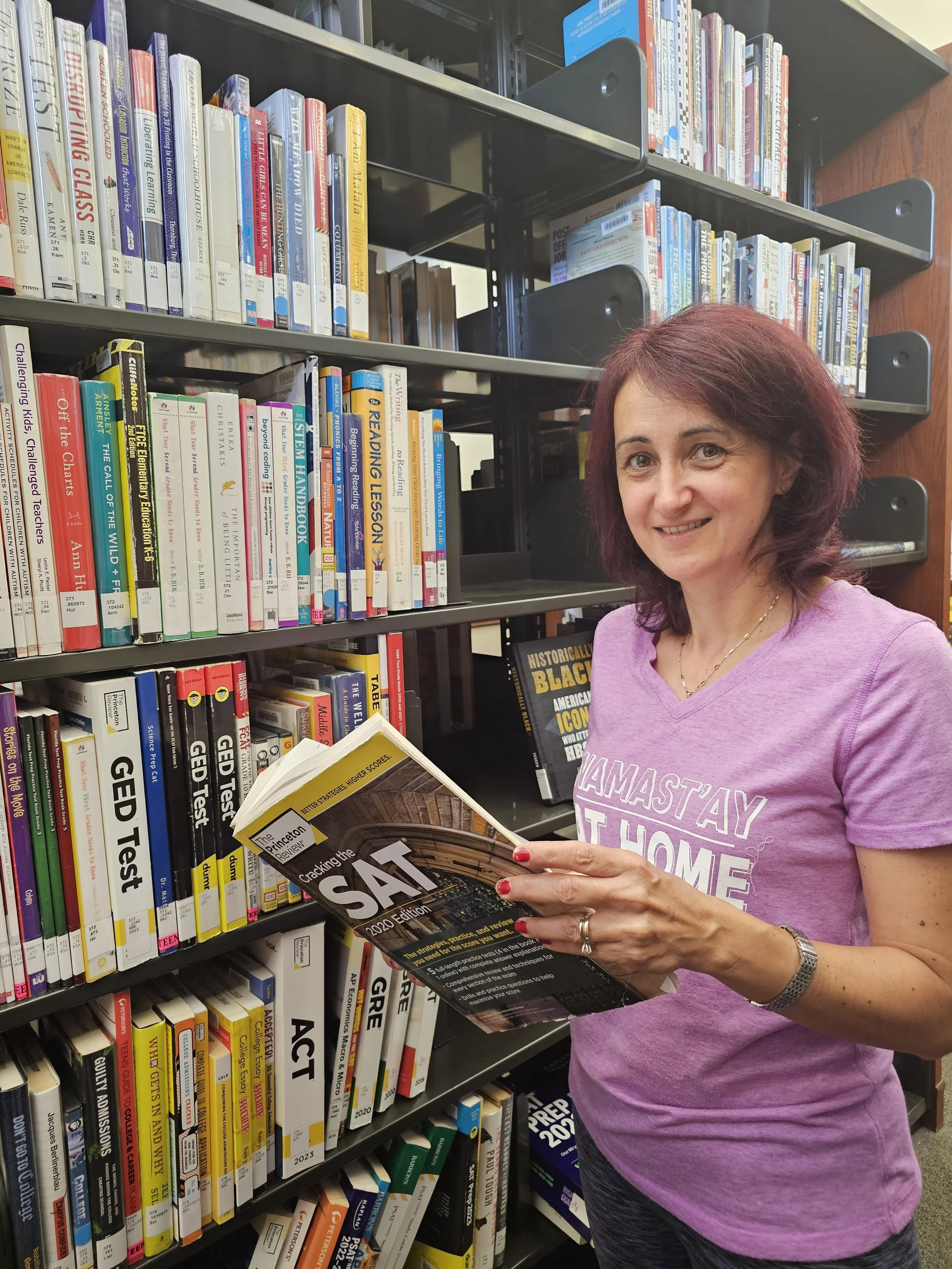 A woman with shoulder-length red hair wearing a purple T-shirt, holding a SAT prep book, standing in a library aisle with bookshelves filled with educational books.