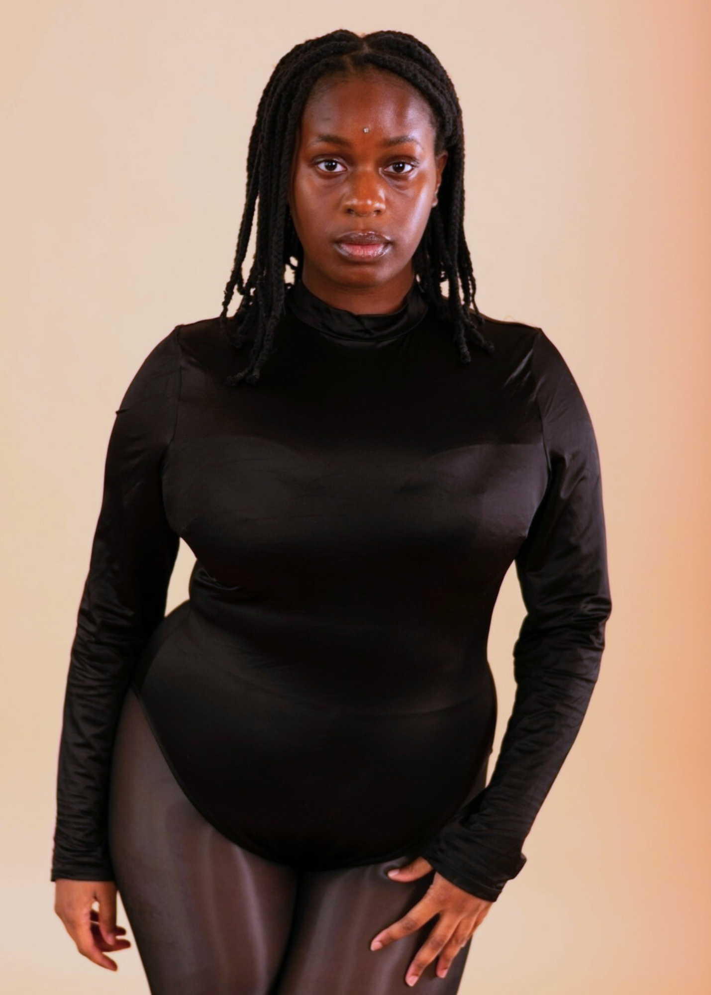A woman with natural dreadlocks wearing a black long-sleeve top and black shiny pants, standing in front of a beige background.